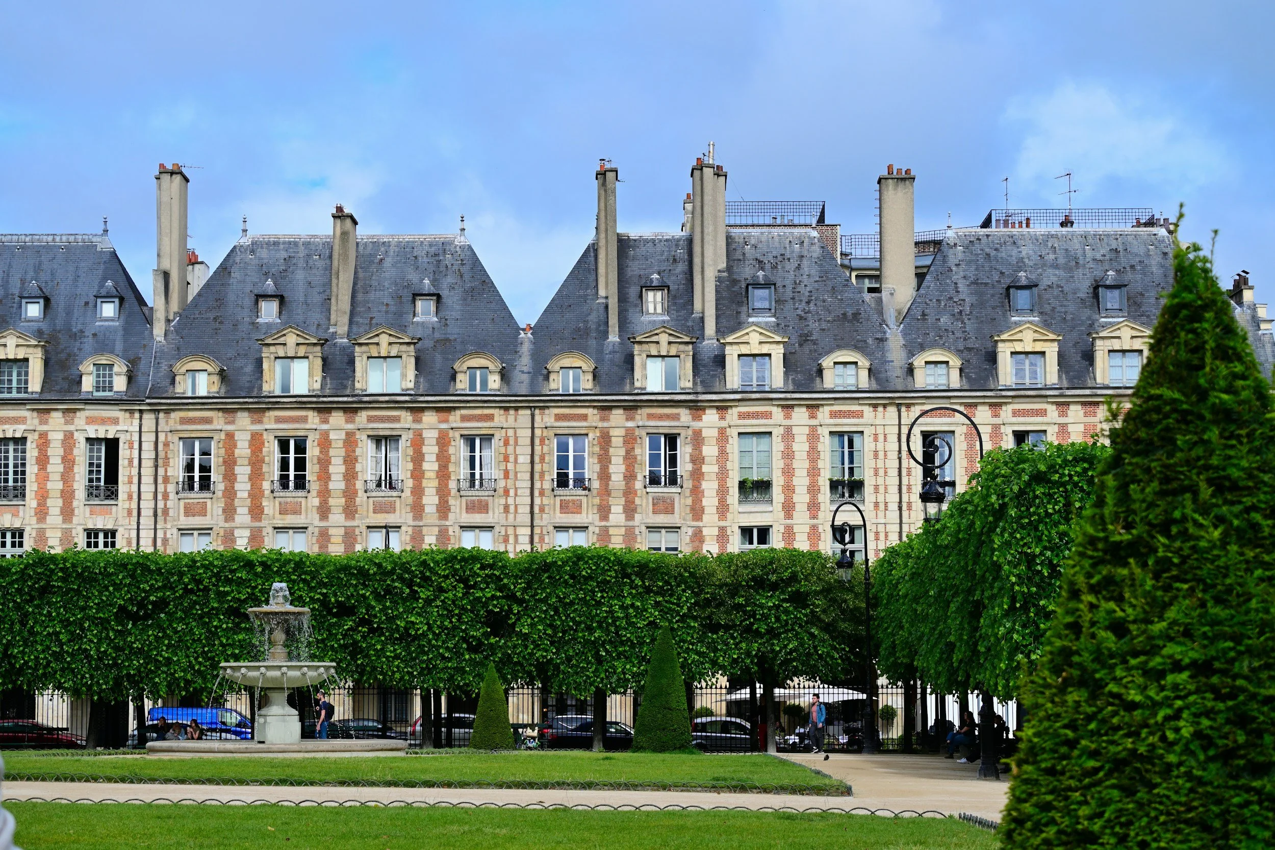 Parisian apartment buildings of La Place des Vosges with classic gray roofs, a park with a fountain, benches, and green trees and bushes in the foreground.