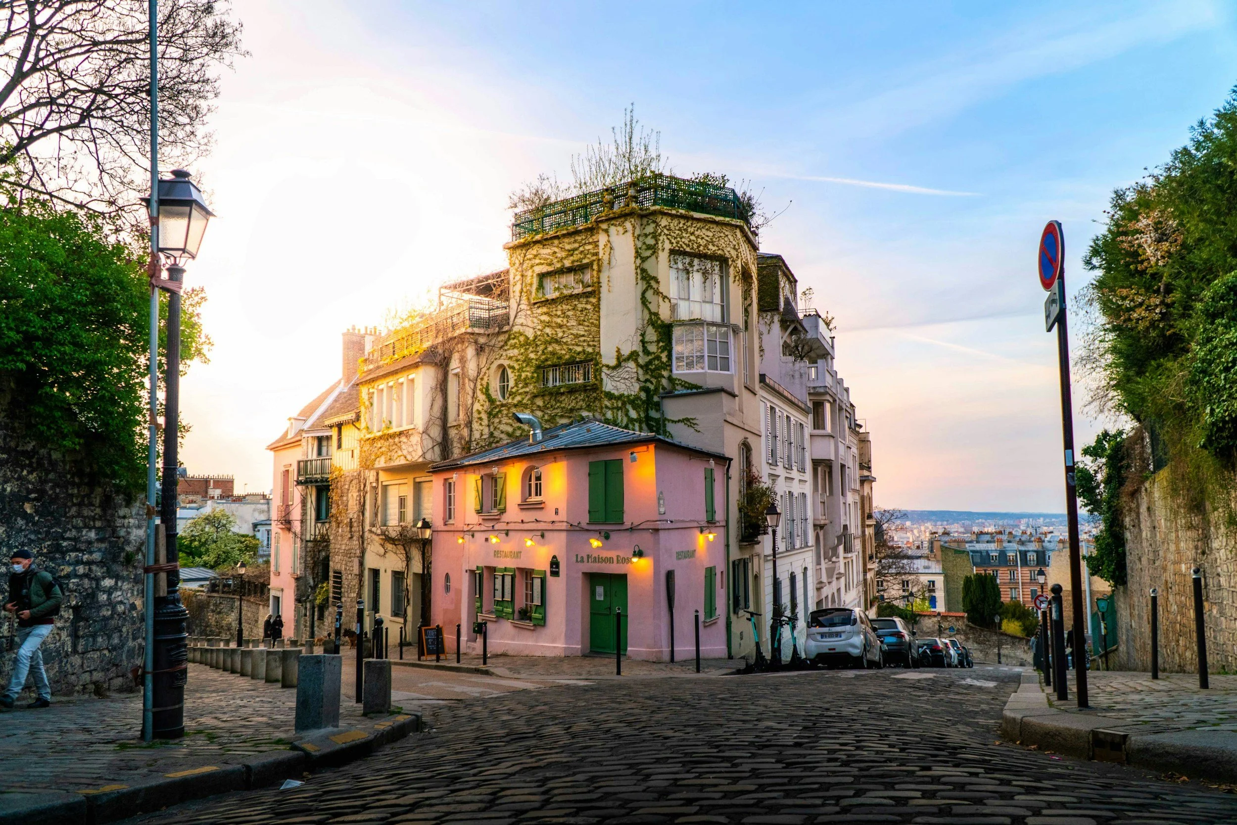 Colorful Parisian street scene of Montmartre neighborhood with cobblestone pavement, charming buildings including a pink building labeled 'La Maison Rose,' and a view of the distant city skyline with a blue sky and light clouds.