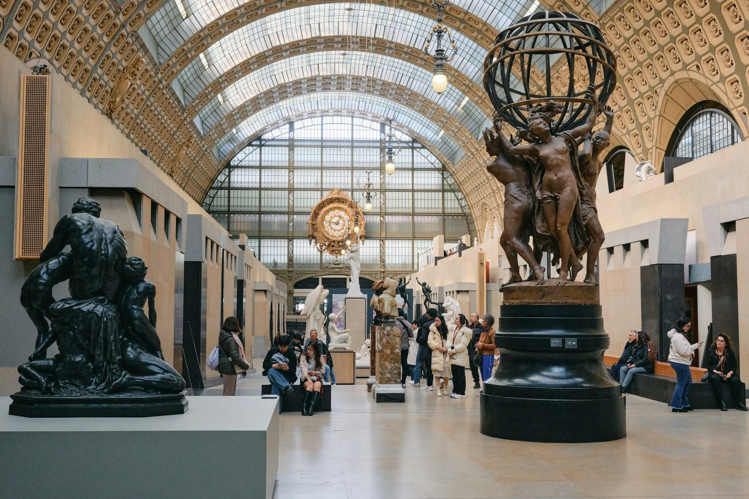 View of Musée d'Orsay with sculptures and visitors, featuring a large glass arched ceiling and ornate clock in the background.