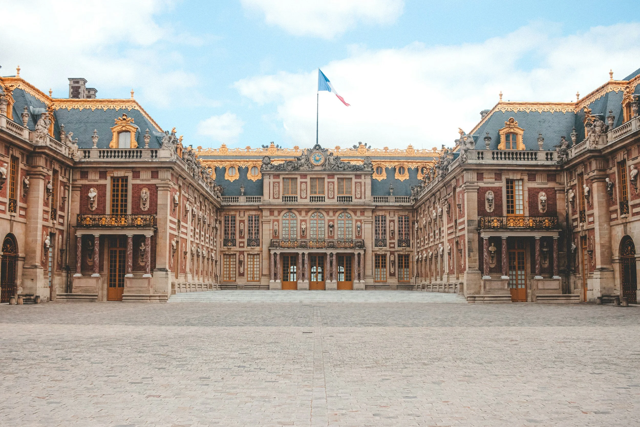 The Palace of Versailles in France under a partly cloudy sky with a French flag flying on top.
