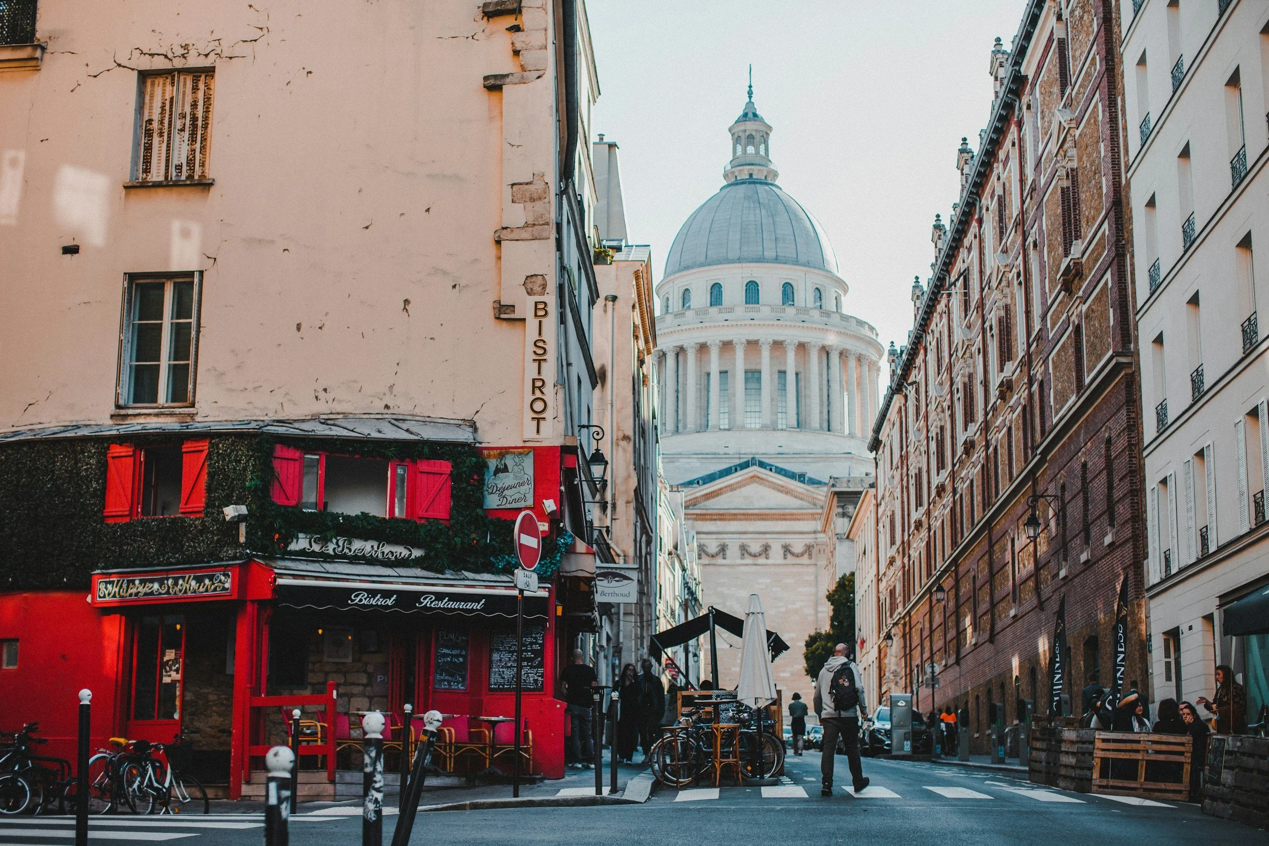 Street scene in Paris with the Pantheon in the background, colorful buildings, pedestrians, bicycles, and a red bistro on the corner.