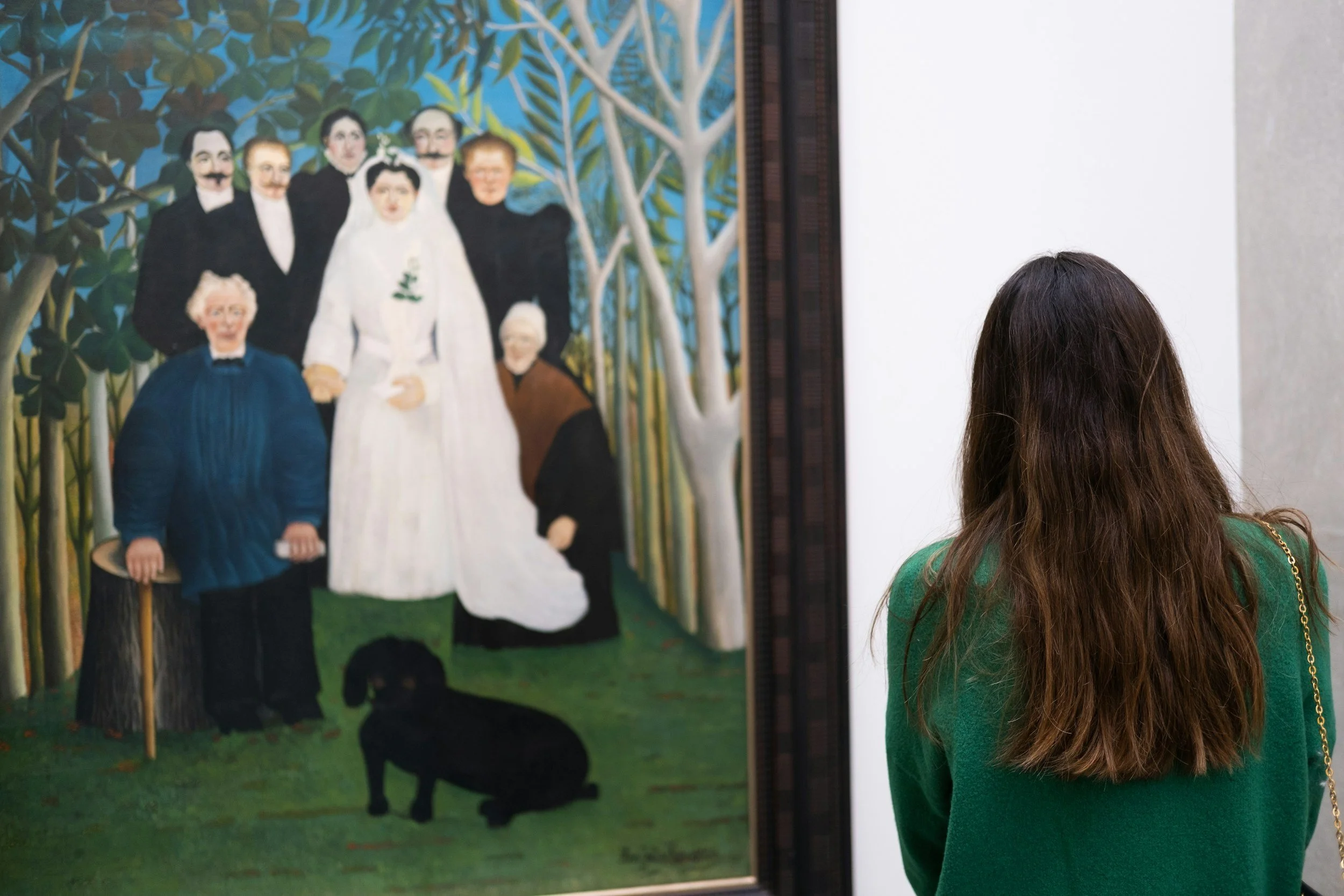A woman with long brown hair wearing a green sweater and a gold chain purse looks at a painting in an art gallery. The painting depicts a group of people outside, including an elderly woman holding a walking stick, a woman in a white dress, a dog, and several men and women standing among trees.