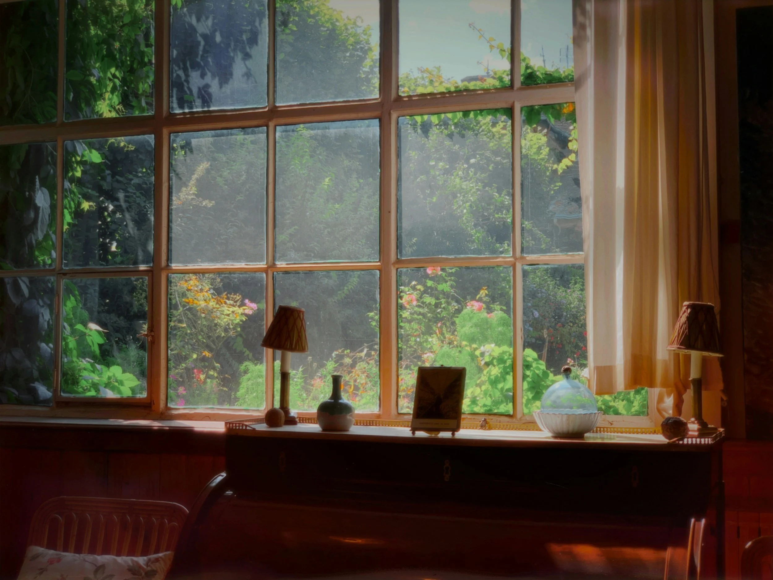 View of the garden outside a large window with multiple panes at Claude Monet's house in Giverny, decorated with small lamps, vases, and a framed photo on the windowsill inside a cozy room.