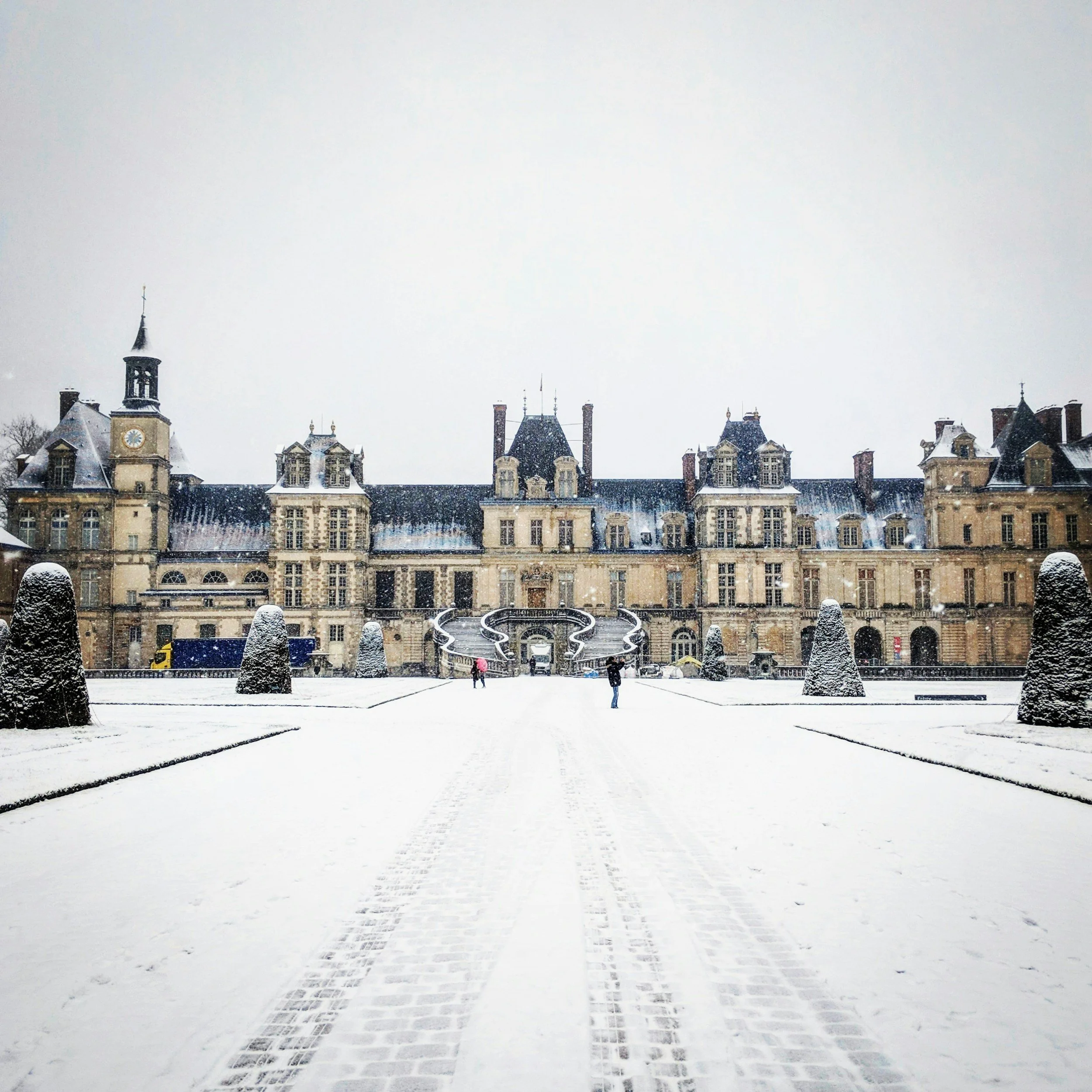 The beautiful castle of Fontainebleau, with a wide snow-covered courtyard and trees covered in snow in front.