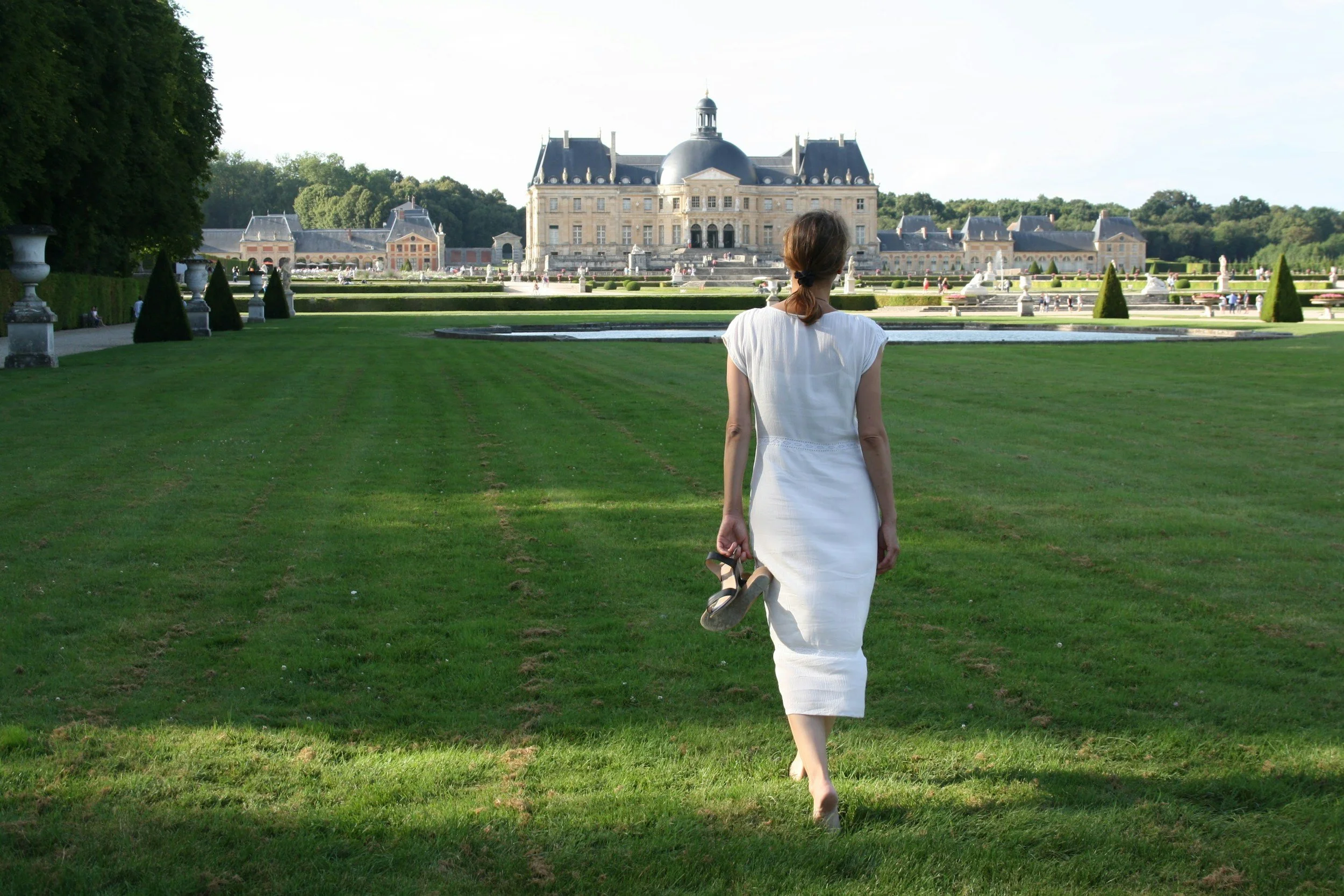 A woman in a white dress walking on a grassy lawn towardsVaux-le-Vicomte palace with fountains and manicured gardens.
