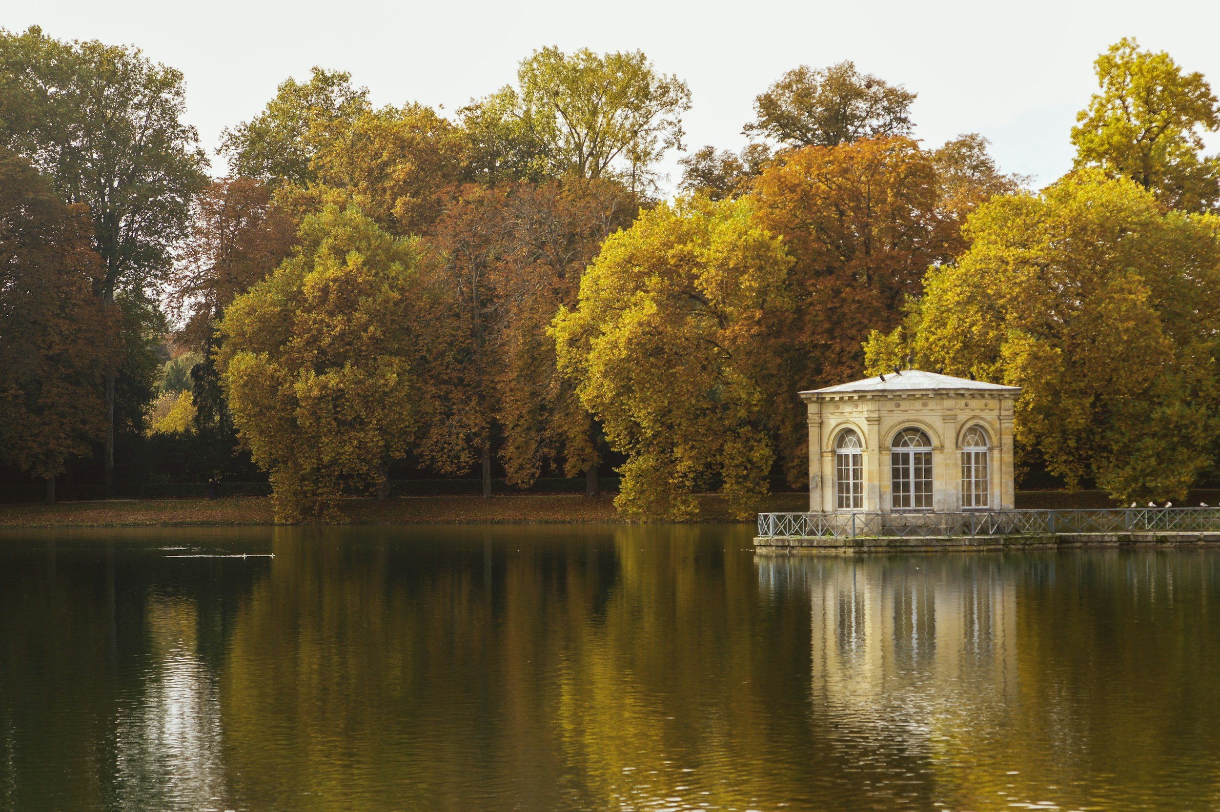 A small stone pavilion with arched windows is on the edge of a calm lake at Fontainebleau Palace, surrounded by trees with autumn leaves in shades of green, yellow, and orange.