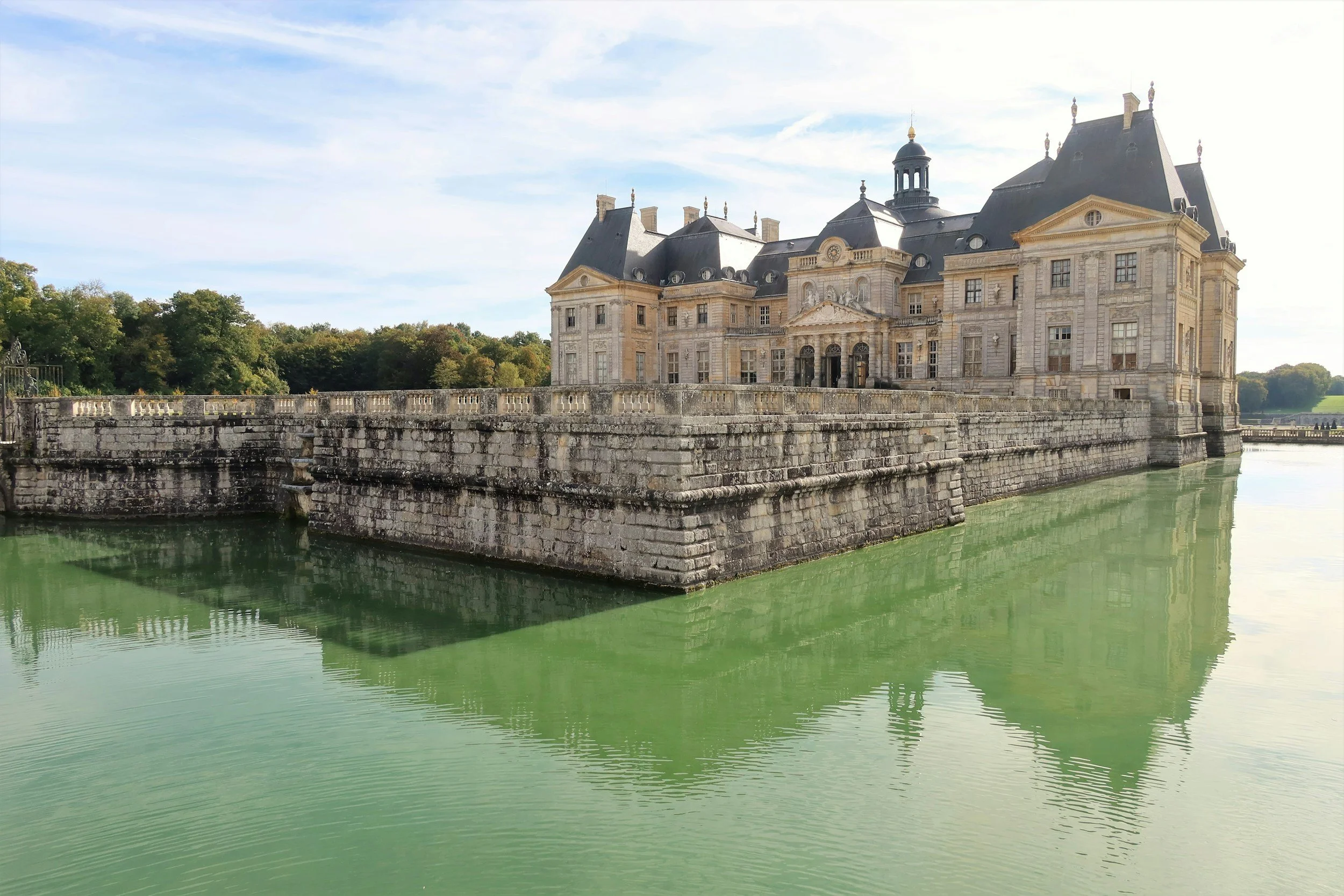 The historic castle of Vaux-le-Vicomte surrounded by a moat filled with green water, with trees in the background and a partly cloudy sky overhead.