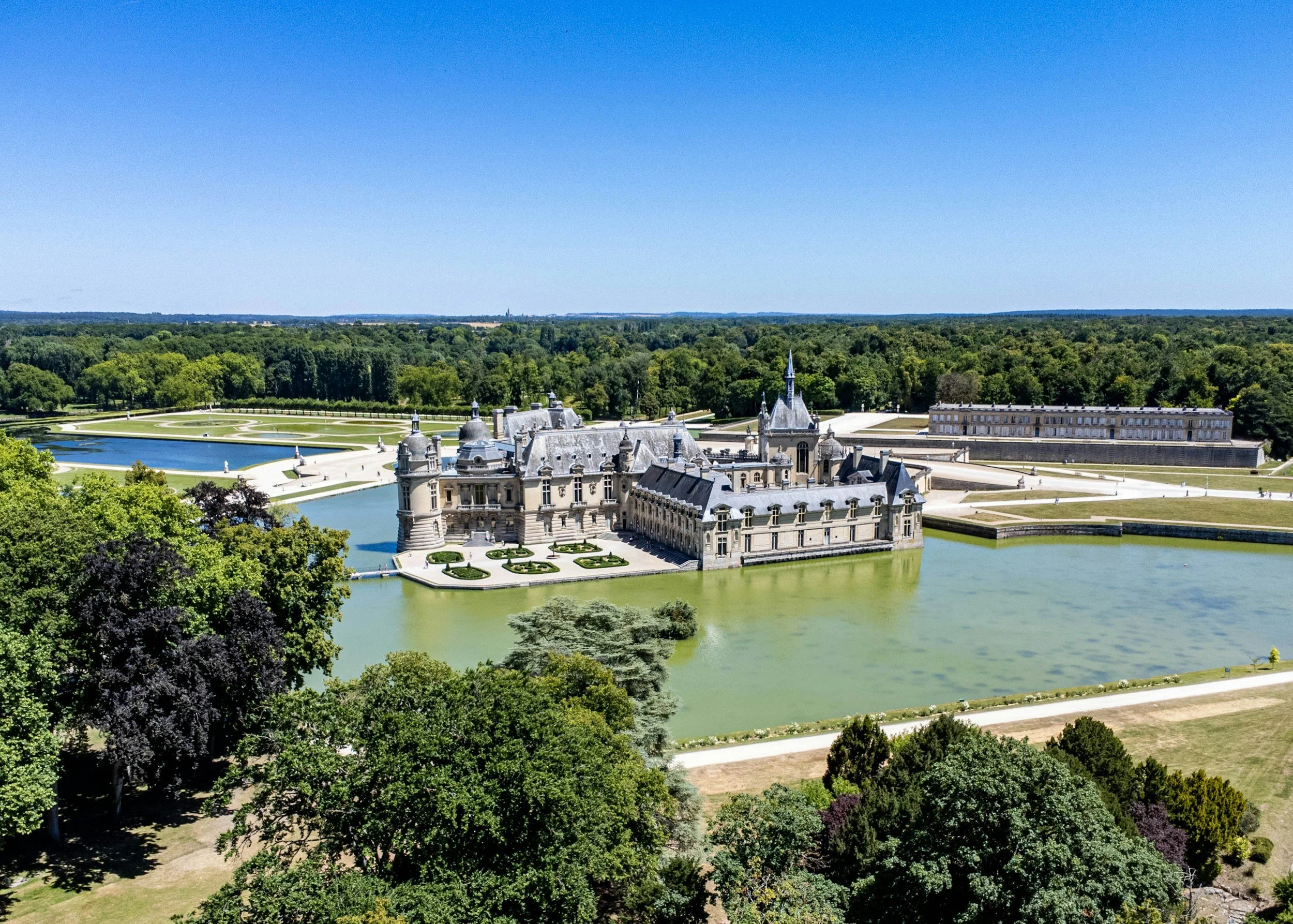 A large historic castle surrounded by water and lush green gardens, with trees in the foreground and a clear blue sky overhead.