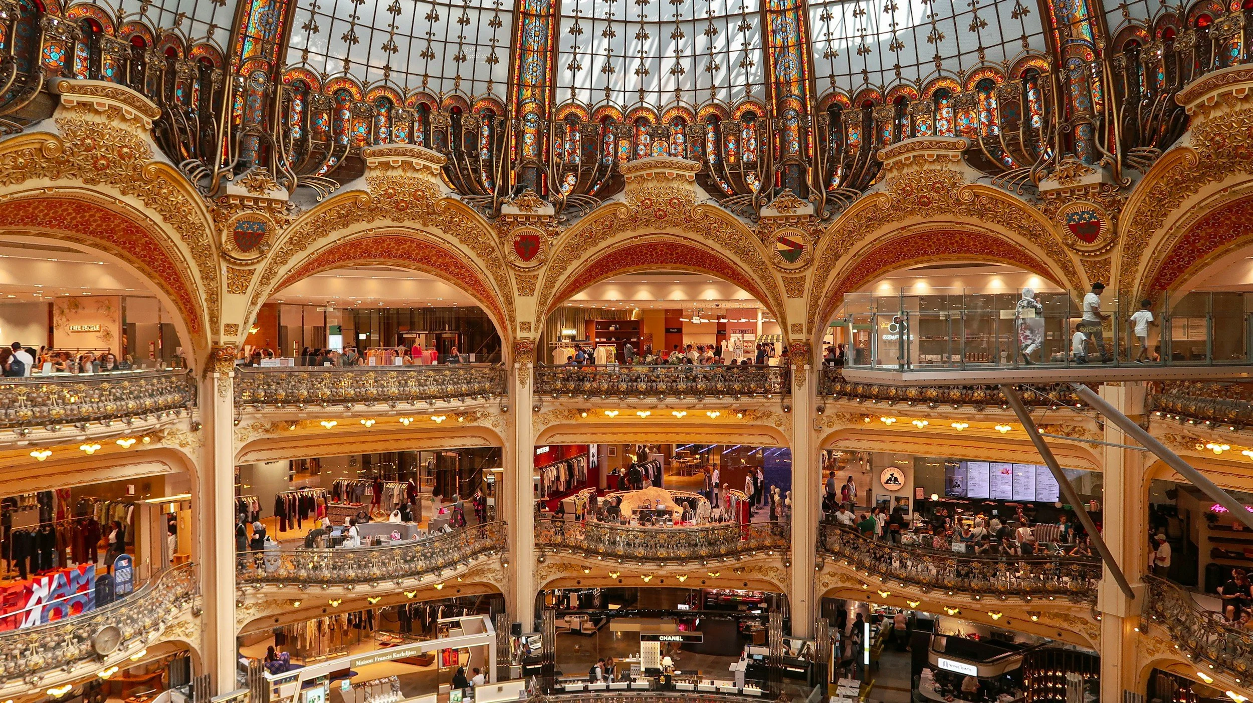 Interior of a luxurious shopping mall with ornate detailed balconies and a large stained glass dome ceiling.