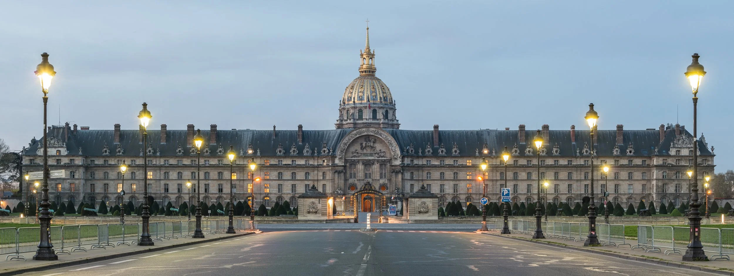 View of the Hôtel des Invalides, a historic building in Paris, France, with its ornate architecture, multiple windows, and street lamps lining the foreground.