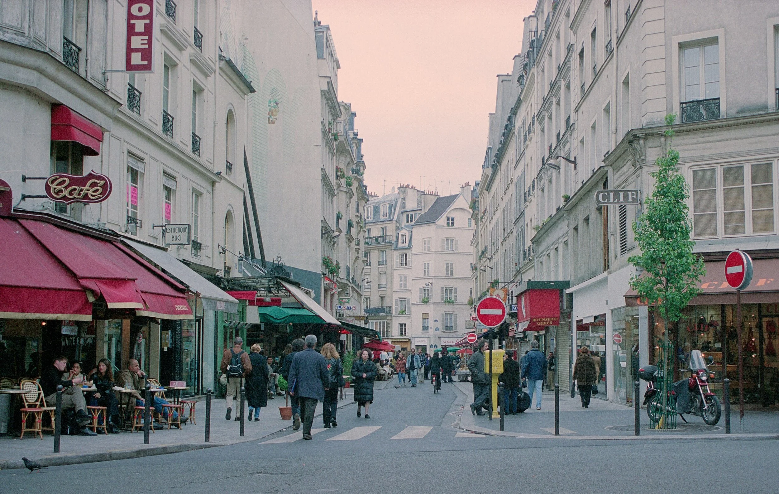 A street scene in Saint-Germain-des-Près area in Paris (France), with people walking on the sidewalk, cafes with red awnings, and white buildings with balconies. Signs indicate cafes and shops, and there is a no-entry traffic sign.