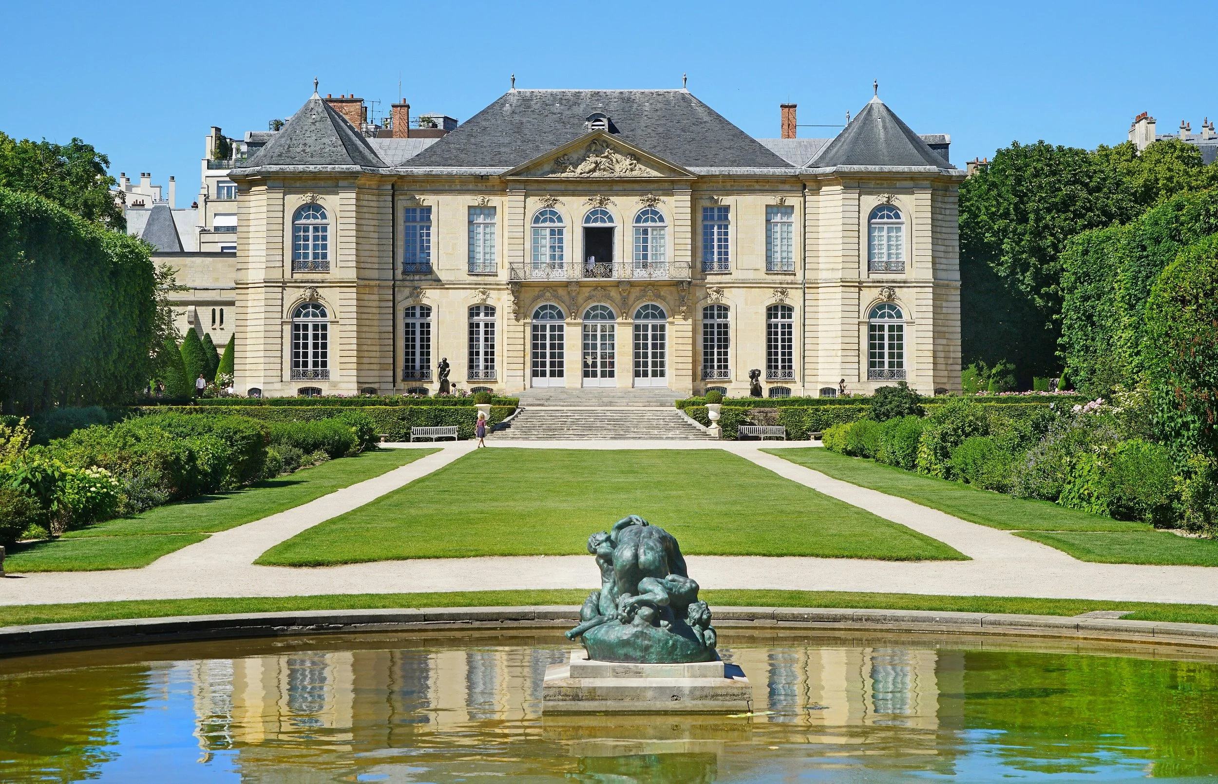 A grand historic mansion with classical architecture, large arched windows, and a slate roof. In the foreground, a sculpture of a reclining figure is positioned in a pond, surrounded by a well-maintained lawn and garden with pathways.