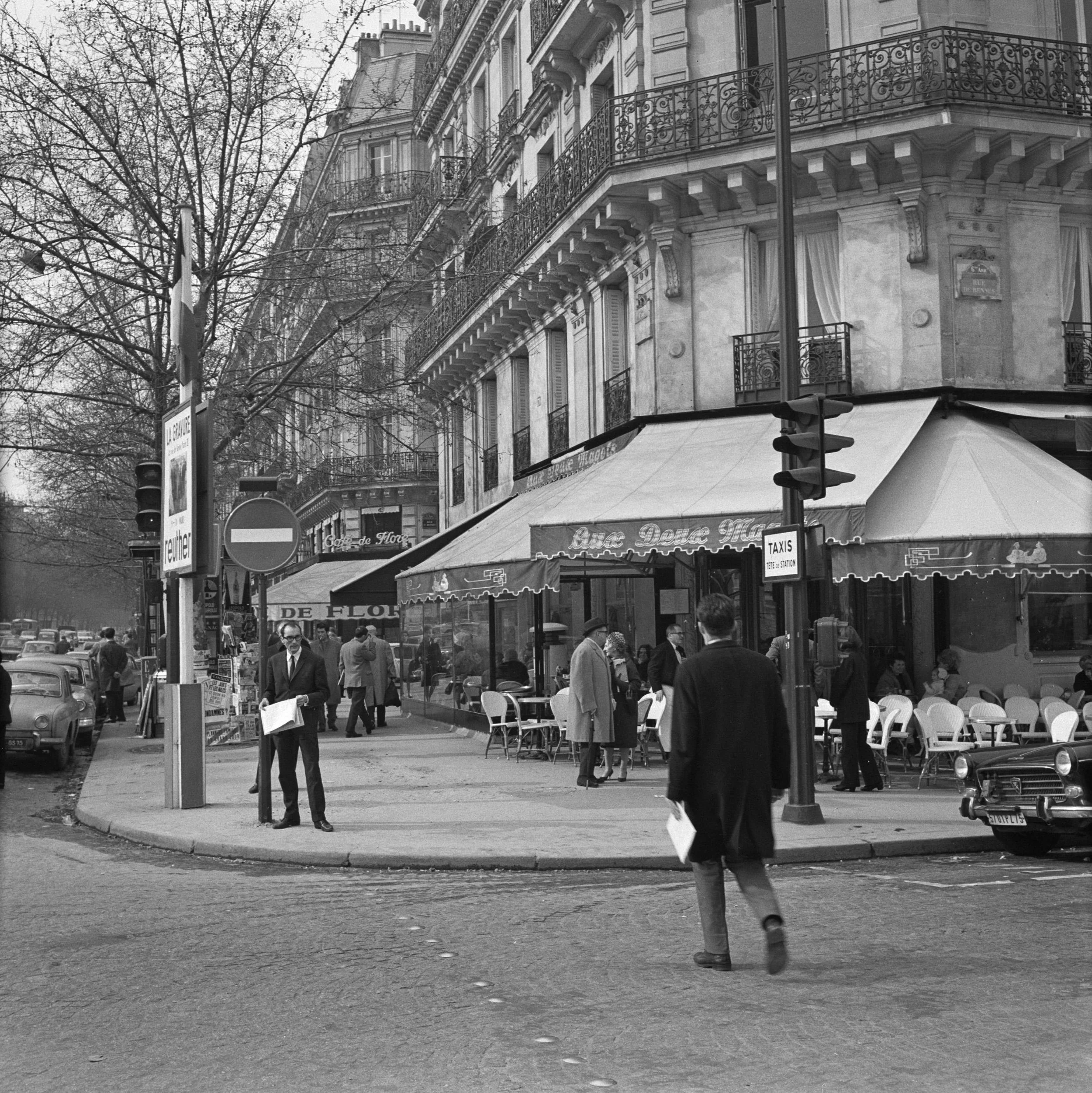 Black-and-white photo of a busy Paris street corner with pedestrians, cars, and a café with outdoor seating under an awning.