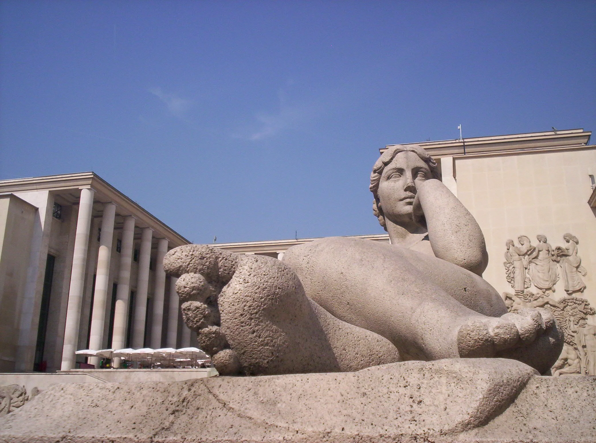 A large stone sculpture of a reclining woman in front of the Musée d'Art Moderne de Paris with one arm resting on her knee and her head propped up by her hand, set against a backdrop of modern classical buildings and a clear blue sky.
