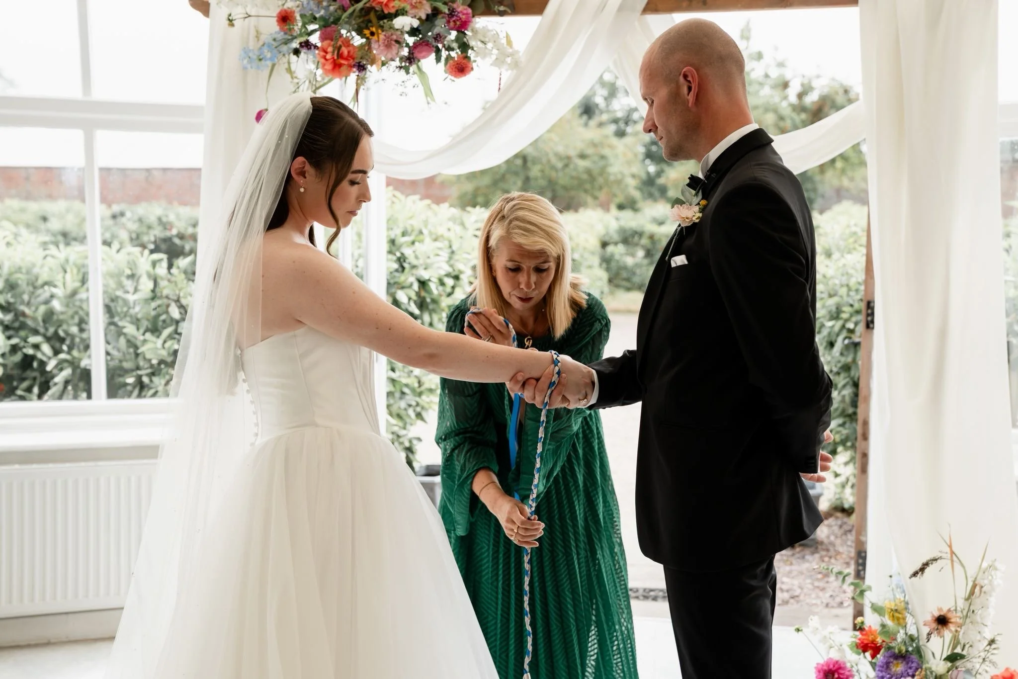 A bride and groom holding hands during their wedding ceremony, with an officiant in green robes reading from a book, under a decorated floral arch in a bright outdoor setting.