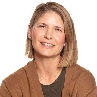 Smiling person with short hair wearing a brown cardigan and black shirt on a white background.