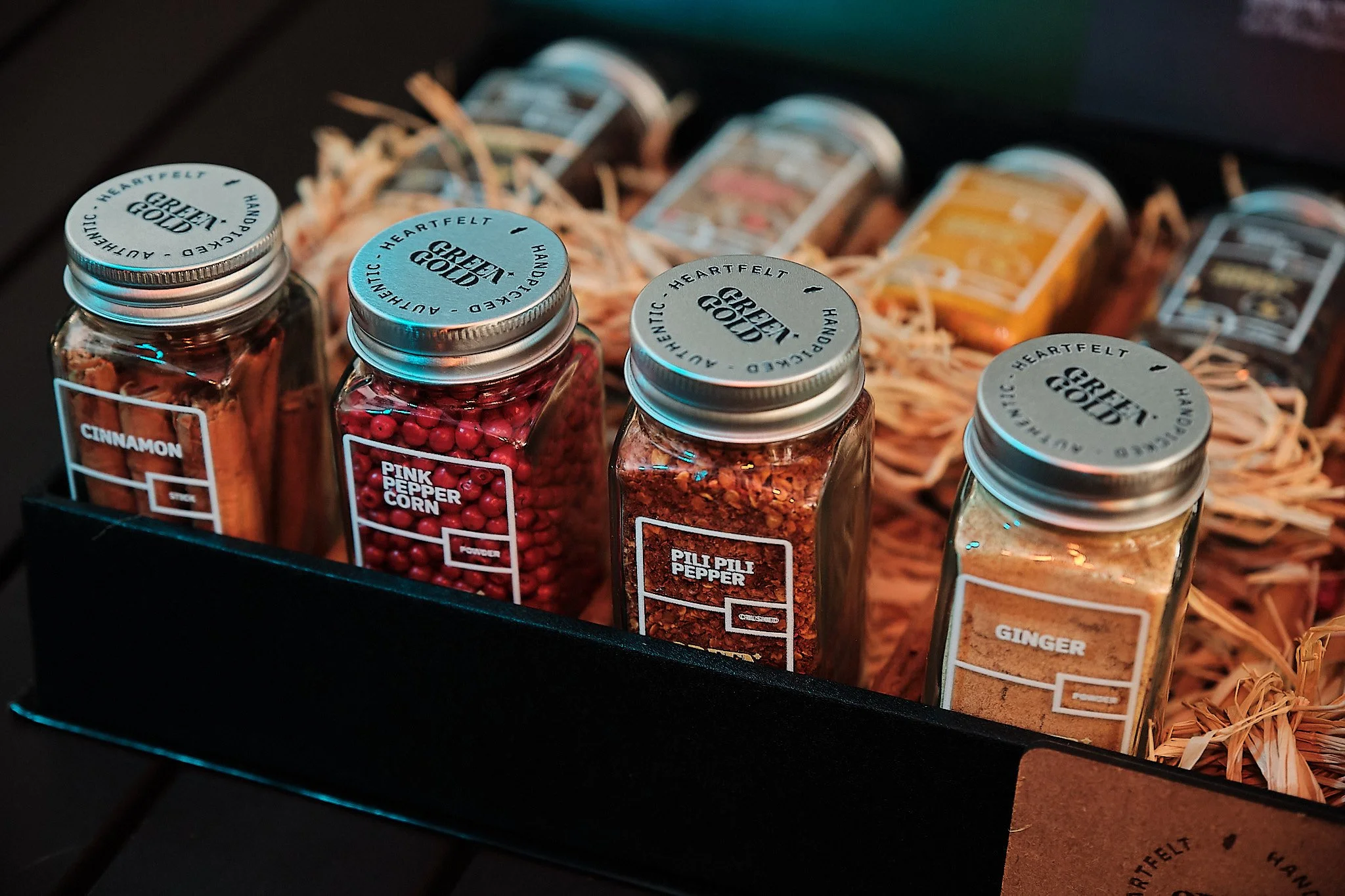 GreenGold - Four glass jars of spices labeled cinnamon, pink peppercorn, pil pil pepper, and ginger, placed inside a black box with shredded paper and other spice jars in the background.