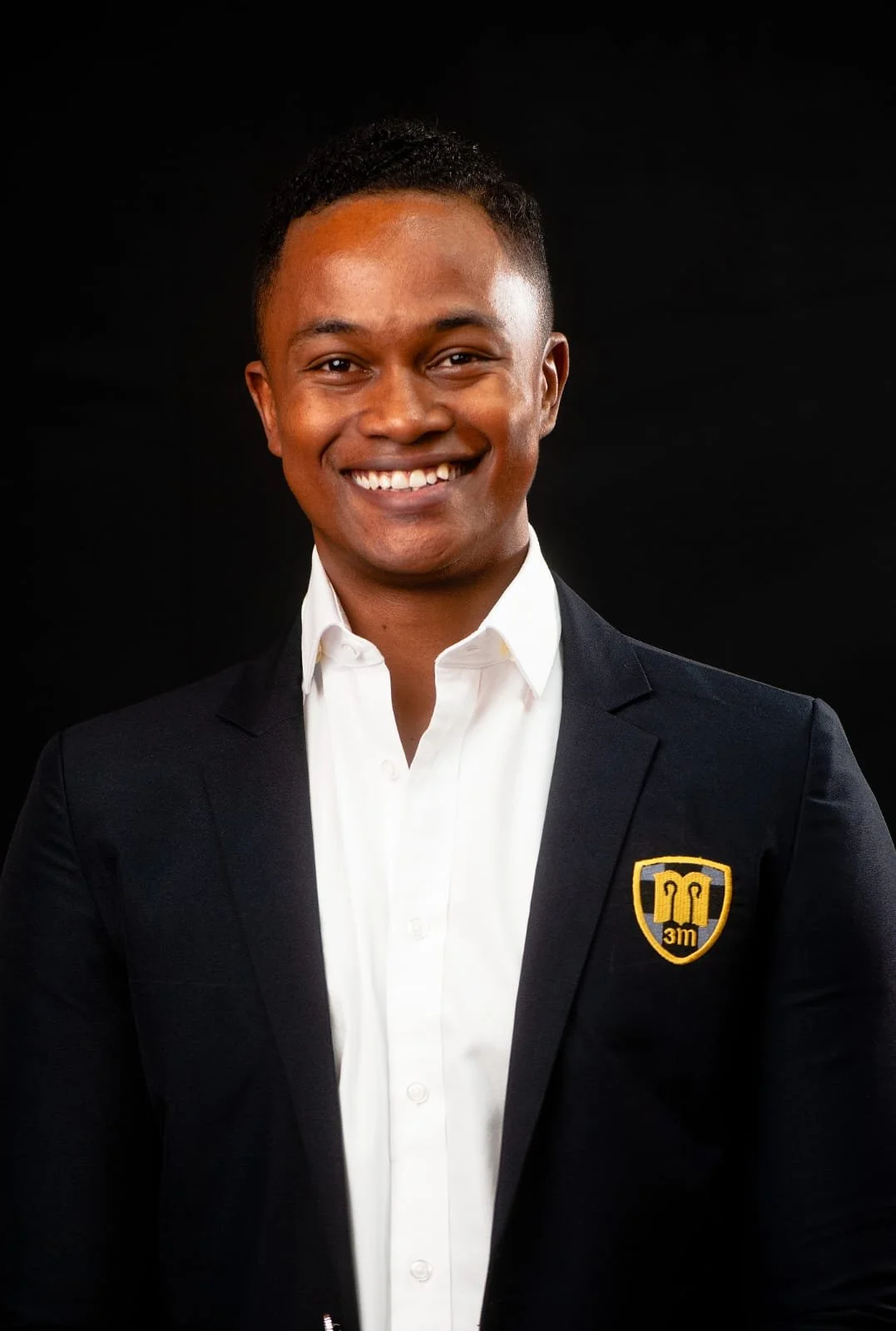 A smiling young man in a suit with a white shirt and a blazer that has a crest with a red shield and a symbol, against a black background.