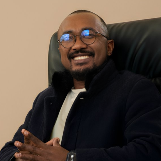 A man with glasses and a beard smiling while sitting on a black leather chair, wearing a dark jacket and a light-colored shirt.