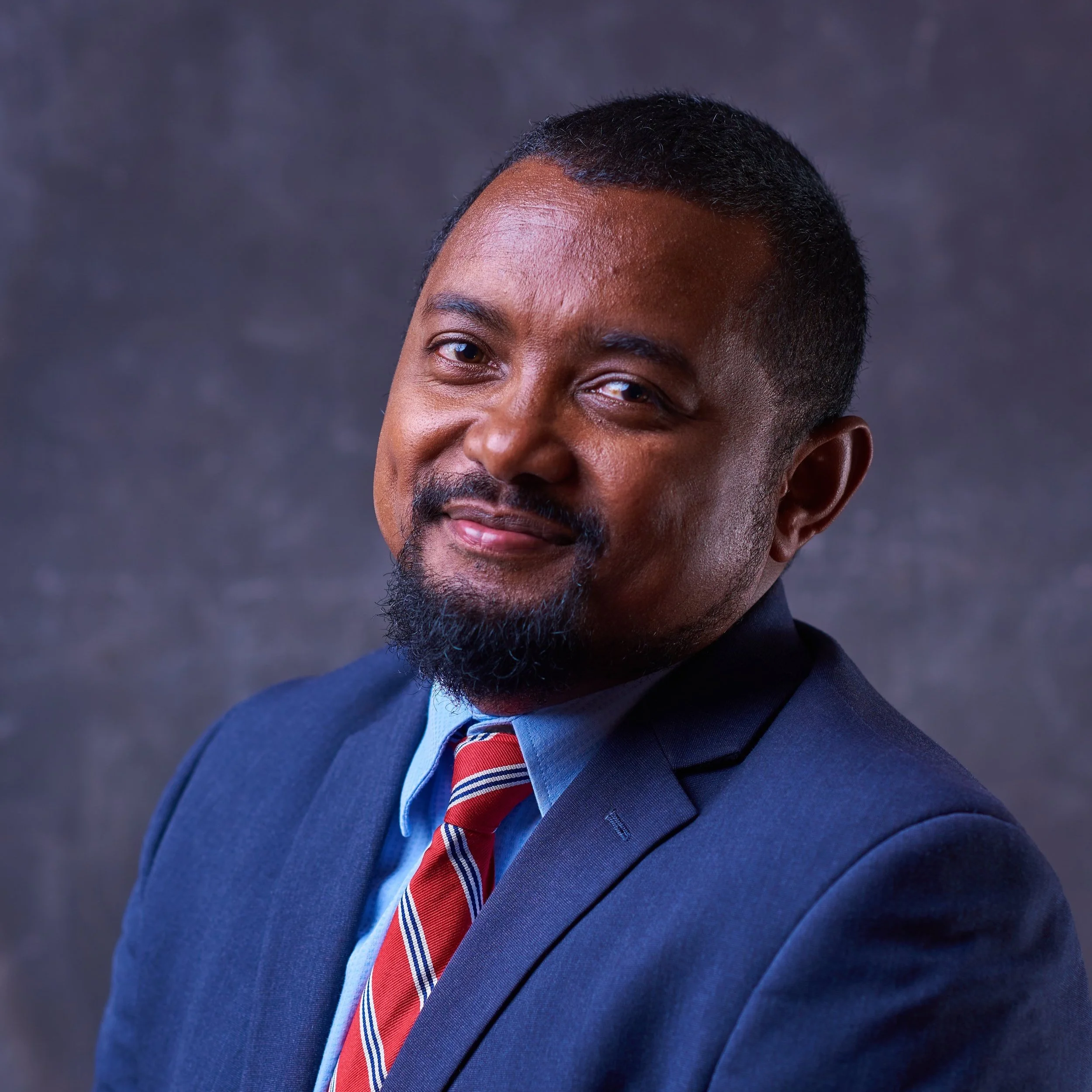 Portrait of a man in a blue suit, light blue shirt, and striped red and blue tie, with short hair and a beard, smiling against a dark grey background.