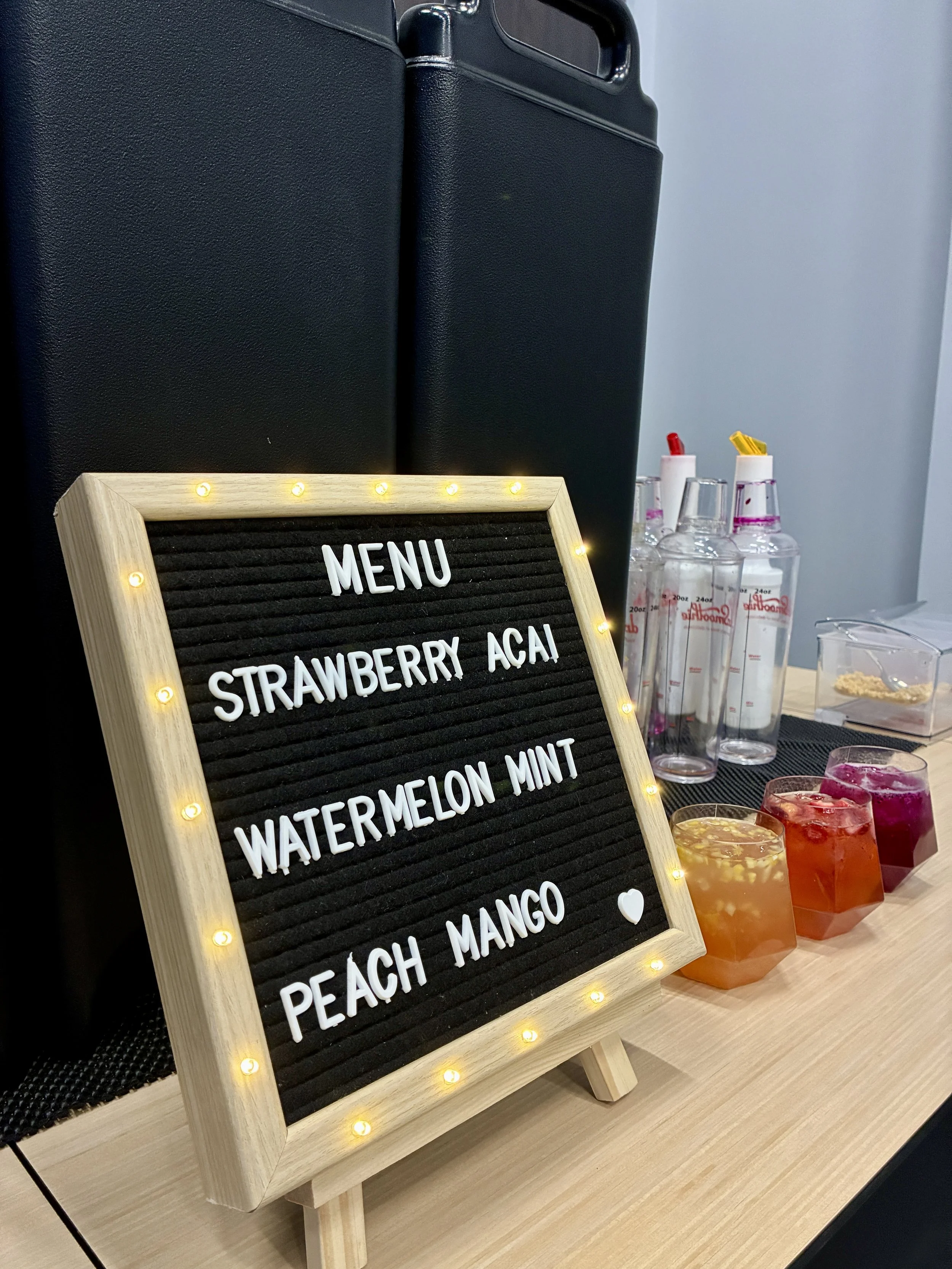 A black board menu with white letters listing flavors: strawberry acai, watermelon mint, peach mango, surrounded by small yellow lights. Behind it are clear bottles with colored caps and straws, and colorful drinks in short cups on a wooden table.