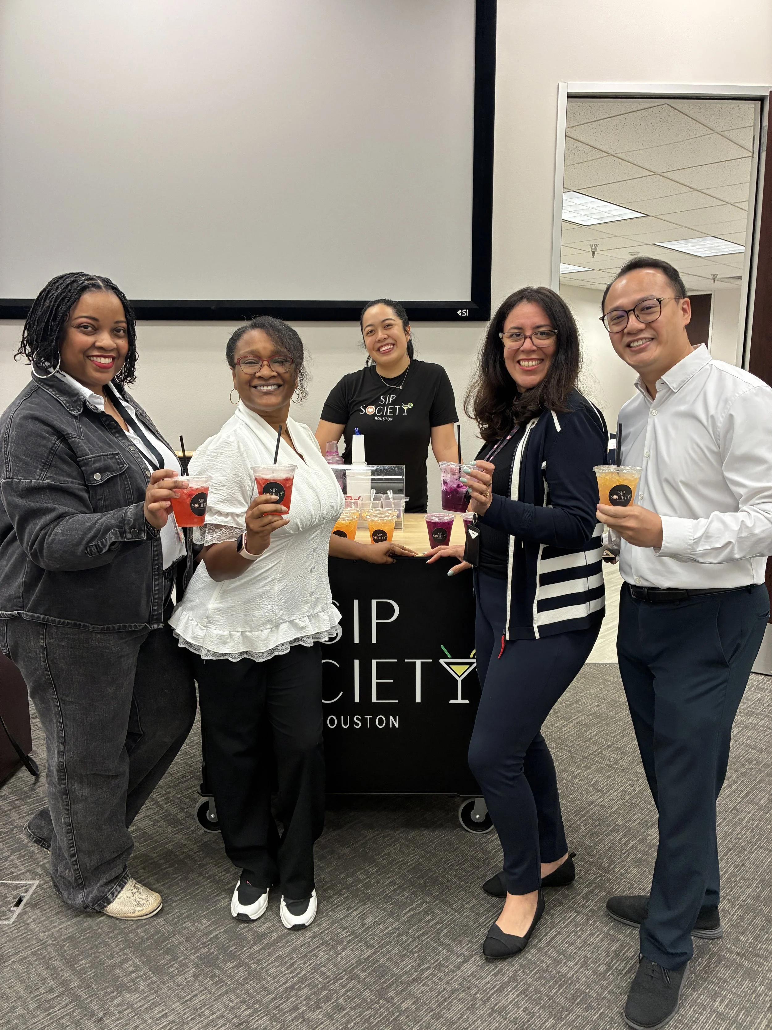 Five diverse people standing together in an office, holding refreshers, smiling, near a mobile refresher cart from Sip Society Houston.
