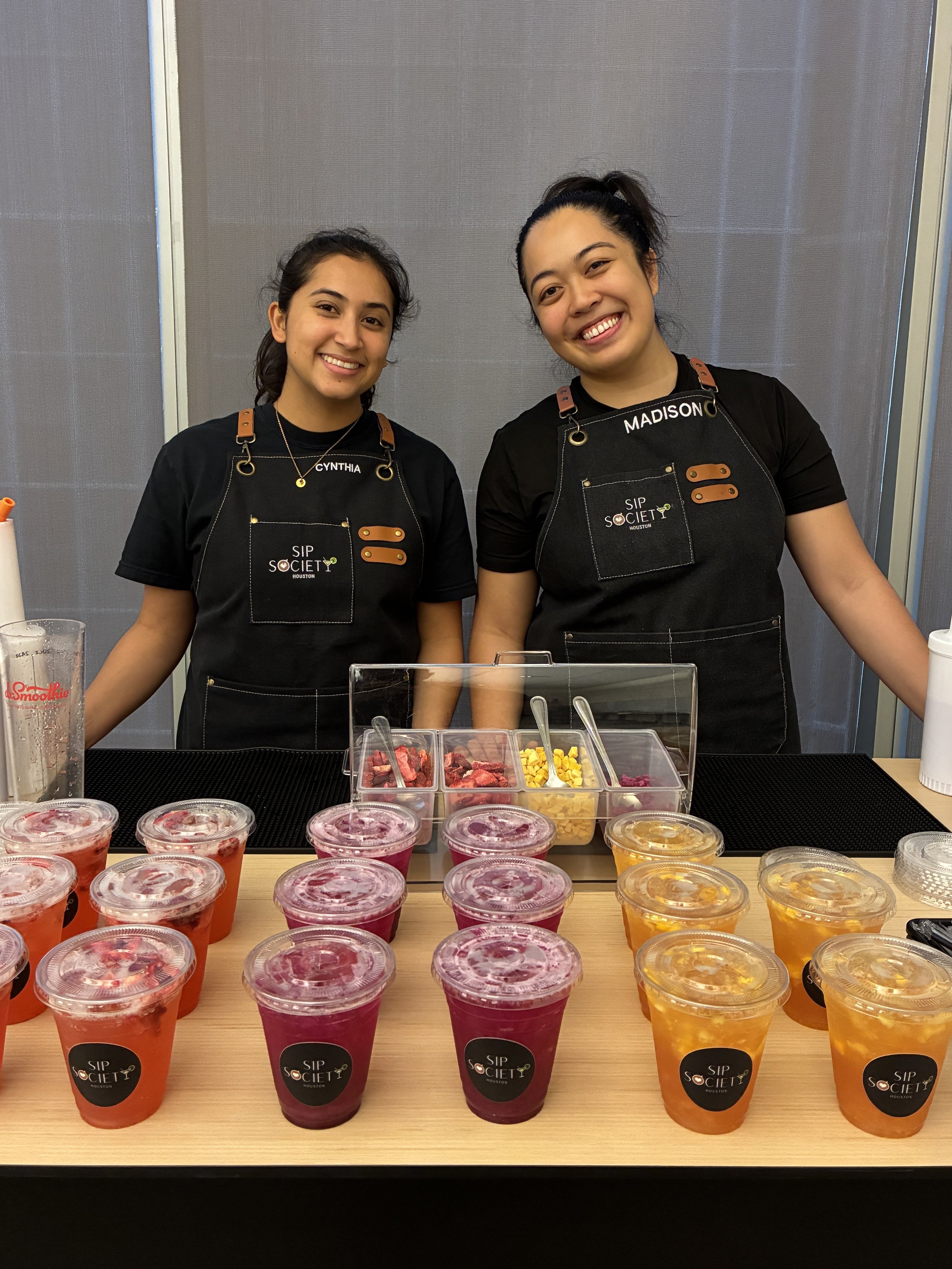 Two women standing behind a table with colorful smoothies in cups, in a store or market setting.