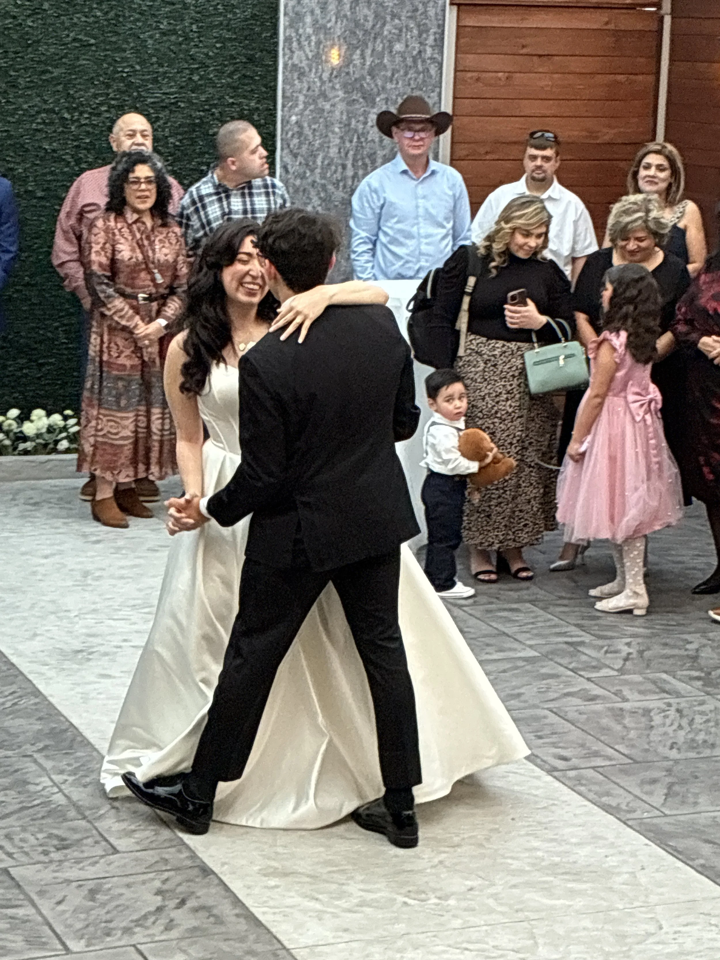 A couple is dancing at a wedding reception, with guests in the background watching and smiling.