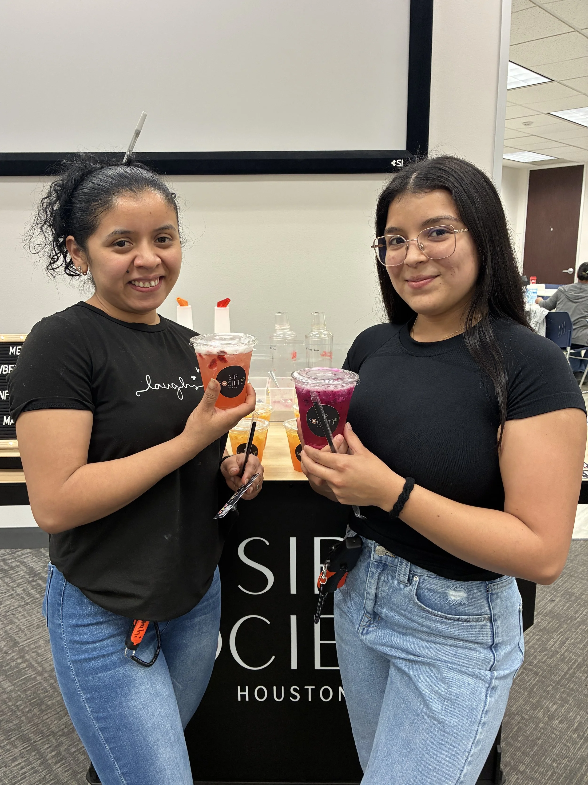 Two young women standing in front of a refresher cart, each holding a colorful refresher provided by Sip Society Houston. One has curly hair tied back, and the other has straight, long hair with glasses.