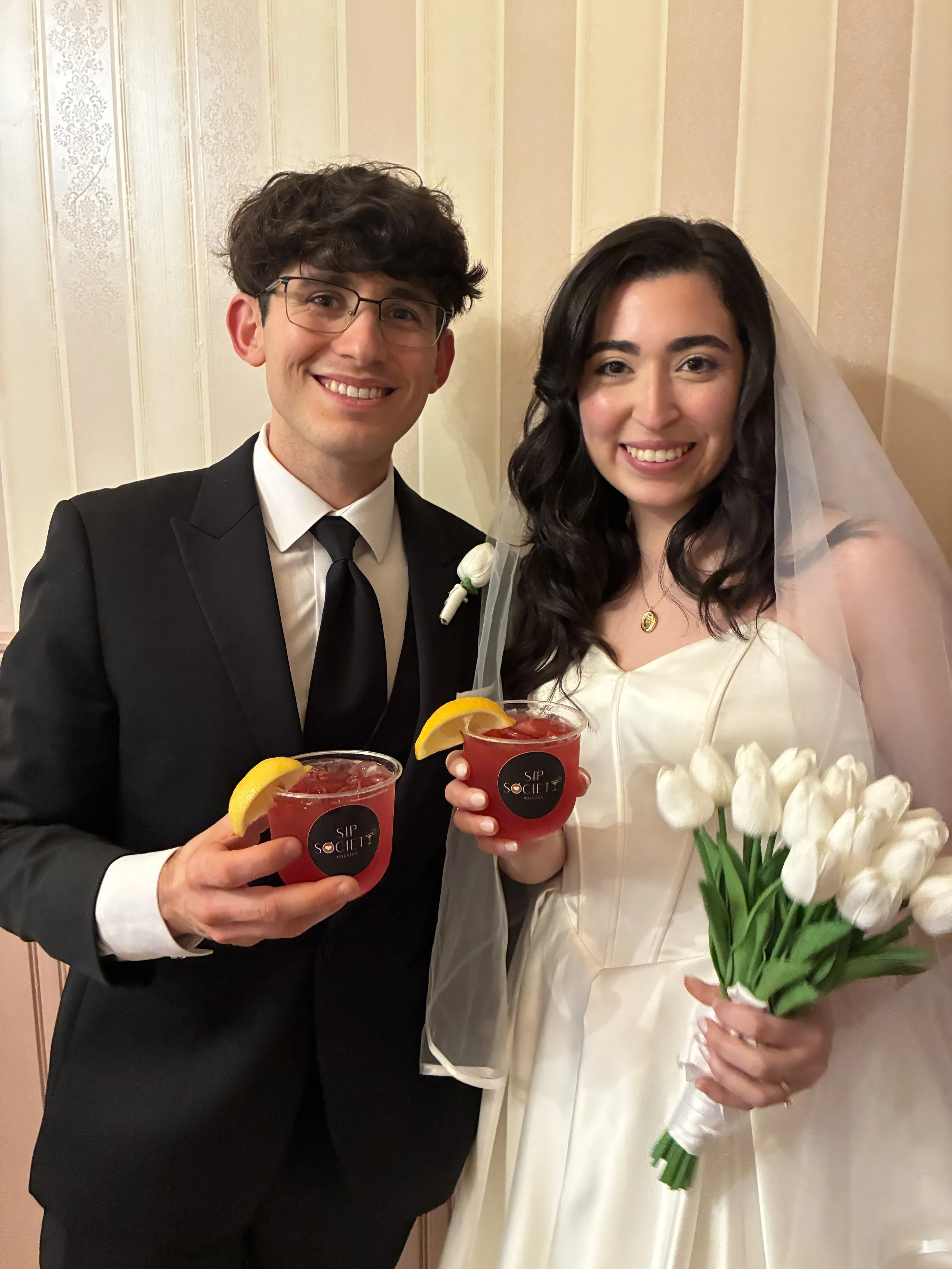 A smiling man in a black tuxedo and a woman in a white wedding dress holding a bouquet of white tulips and Sip Society Houston cocktails, standing indoors against a beige wall.