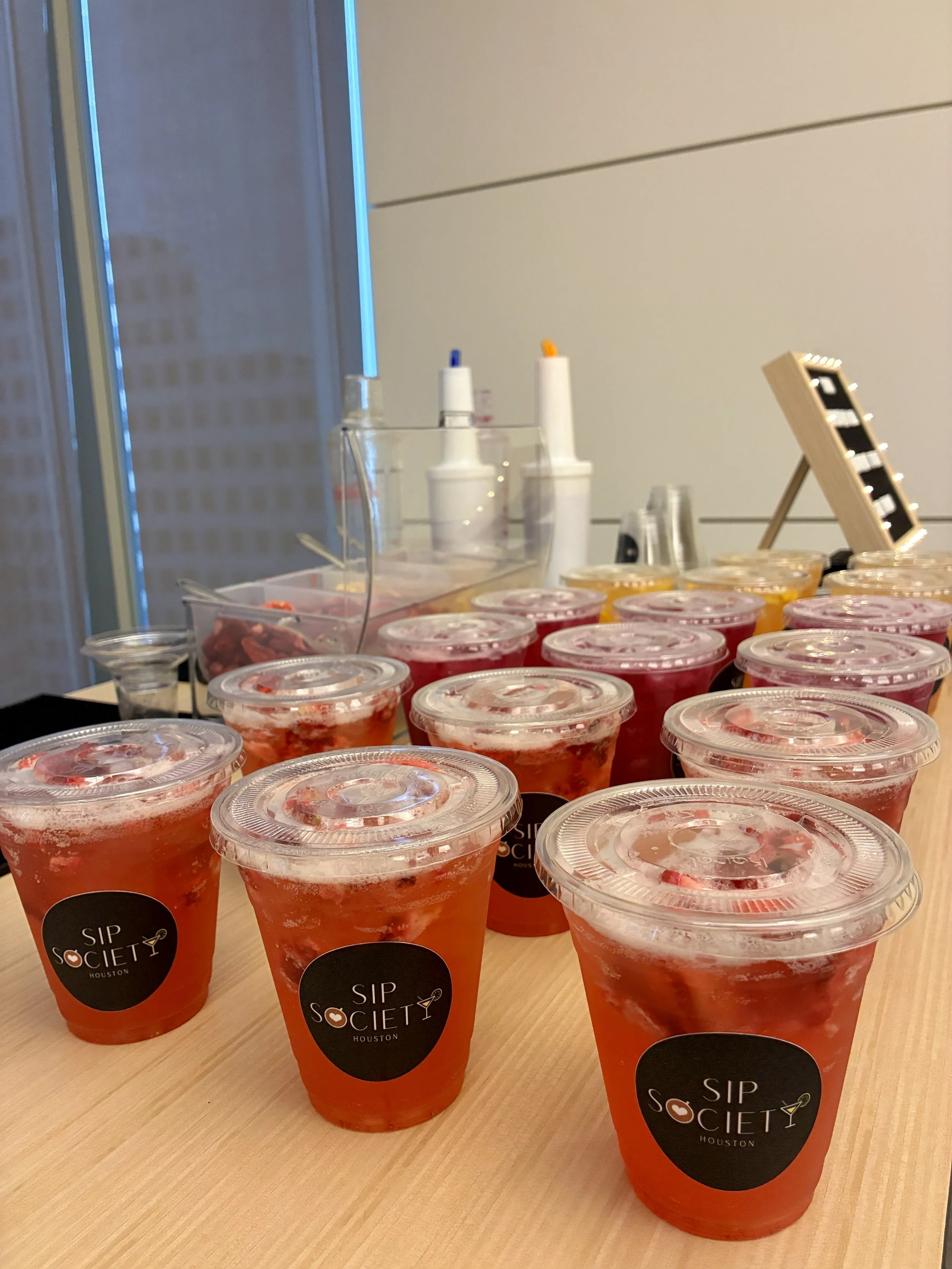 Multiple plastic cups filled with different flavored refreshers, labeled 'Sip Society Houston' on mobile refresher cart.