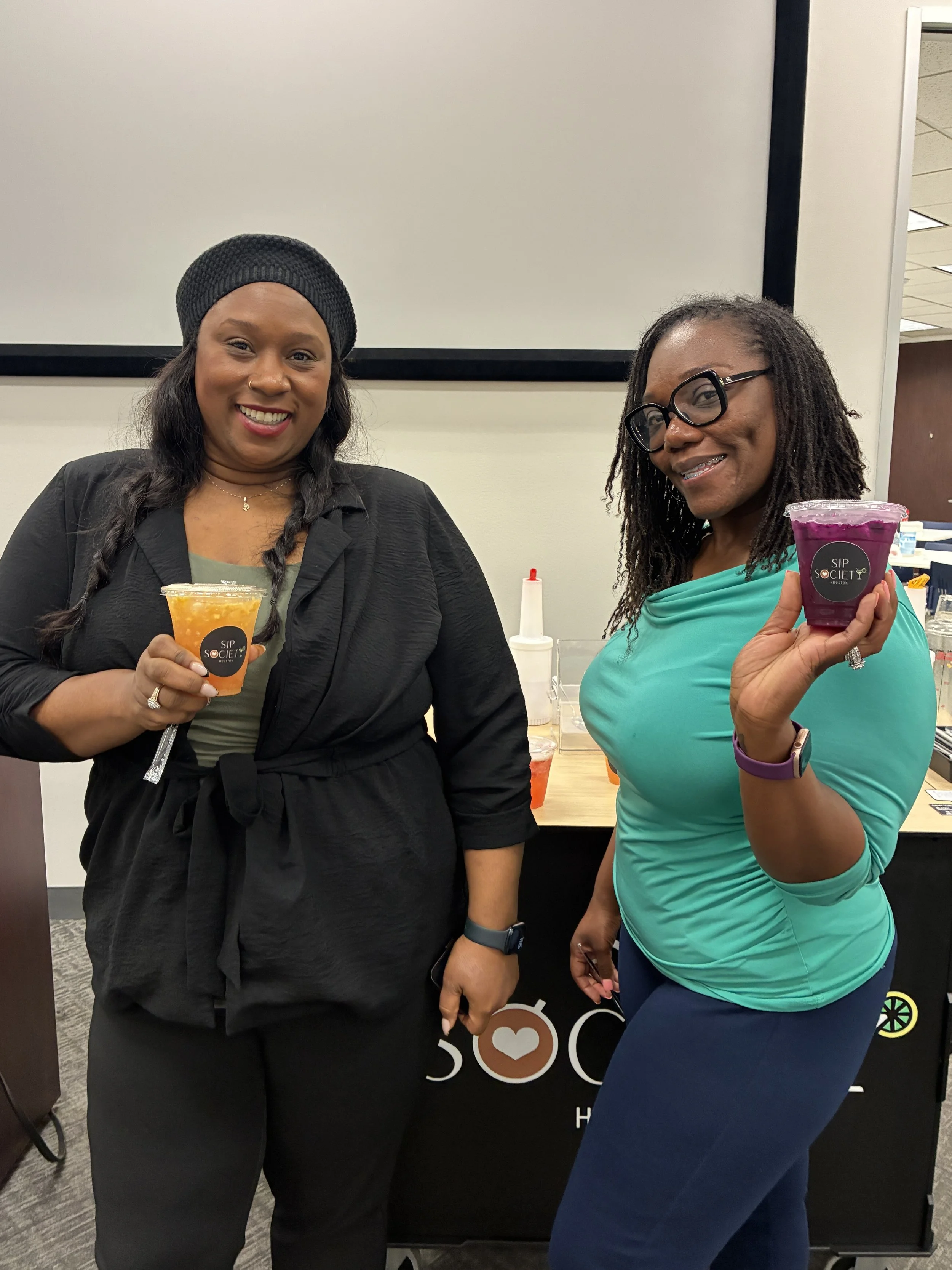 Two women standing indoors, each holding a colorful drink. They are smiling and posing for the photo while holding refreshers from Sip Society Houston.