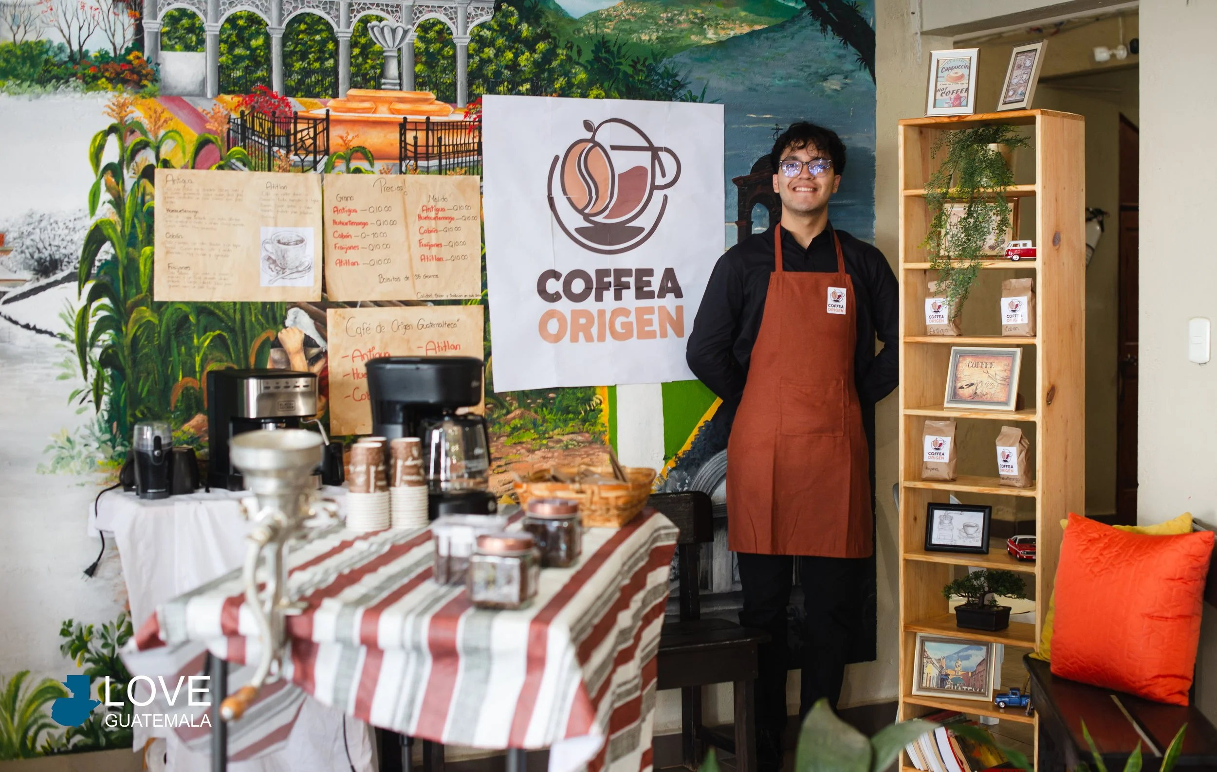 Joven barista sonriente de pie junto a una mesa con máquinas de café y paquetes en una cafetería llamada 'Coffea Origen', paredes decoradas con un mural y estantería con productos y decoraciones, en Guatemala.