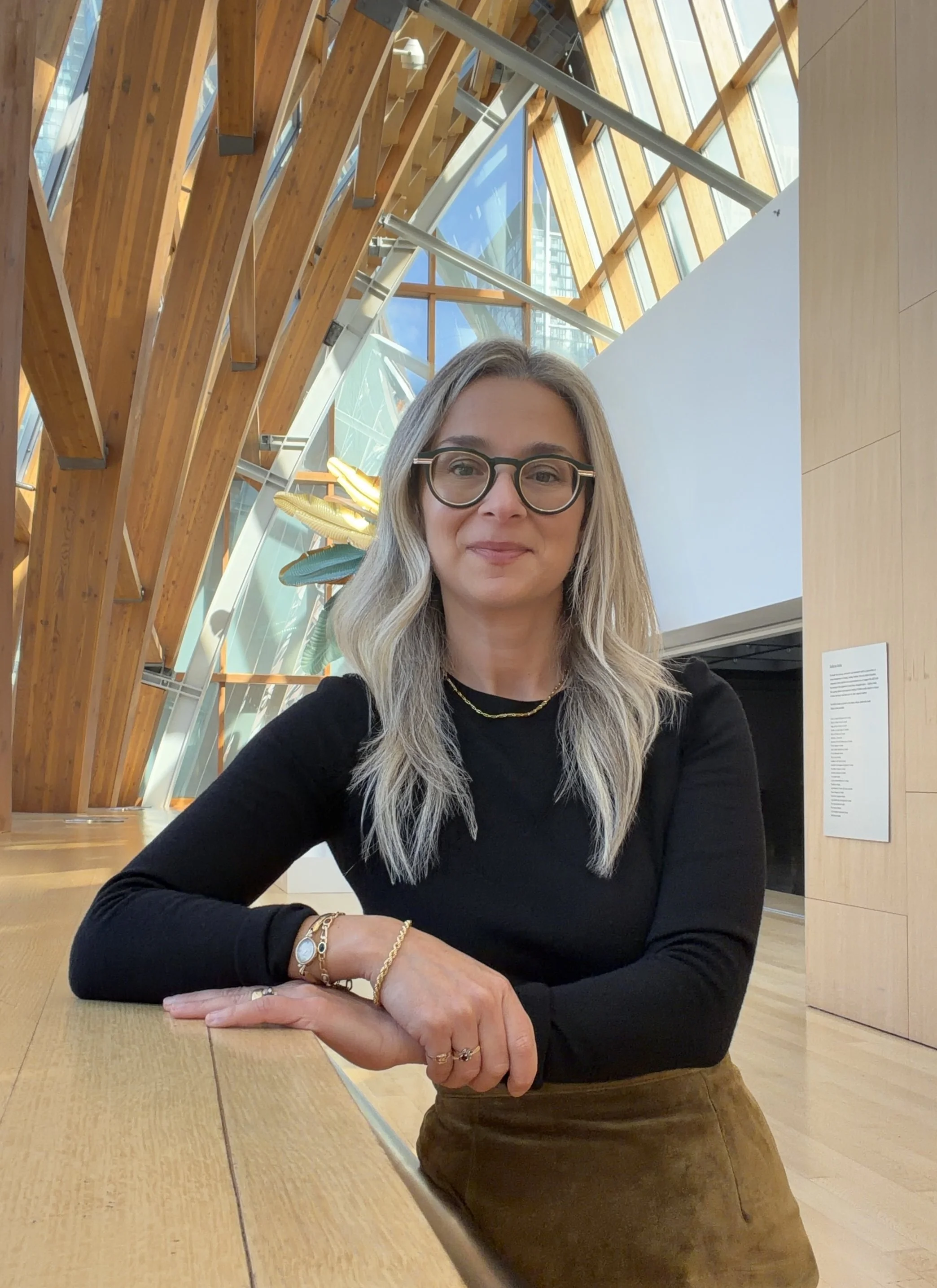 A woman with blonde hair, glasses, and a gold necklace, in a modern indoor space with large windows, wooden beams, and colorful hanging art installations.