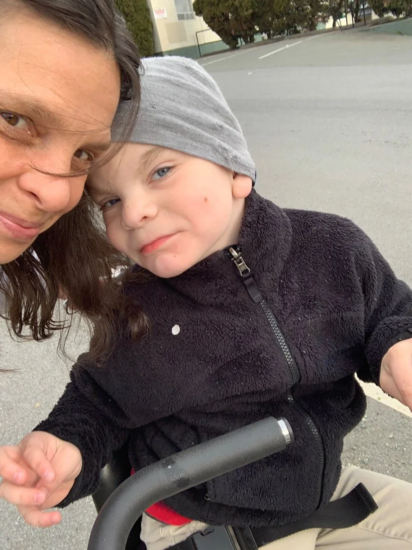 Mother and child smiling with their head touching. Child is sitting in an adaptive stroller while me mother stands and leans into the child.