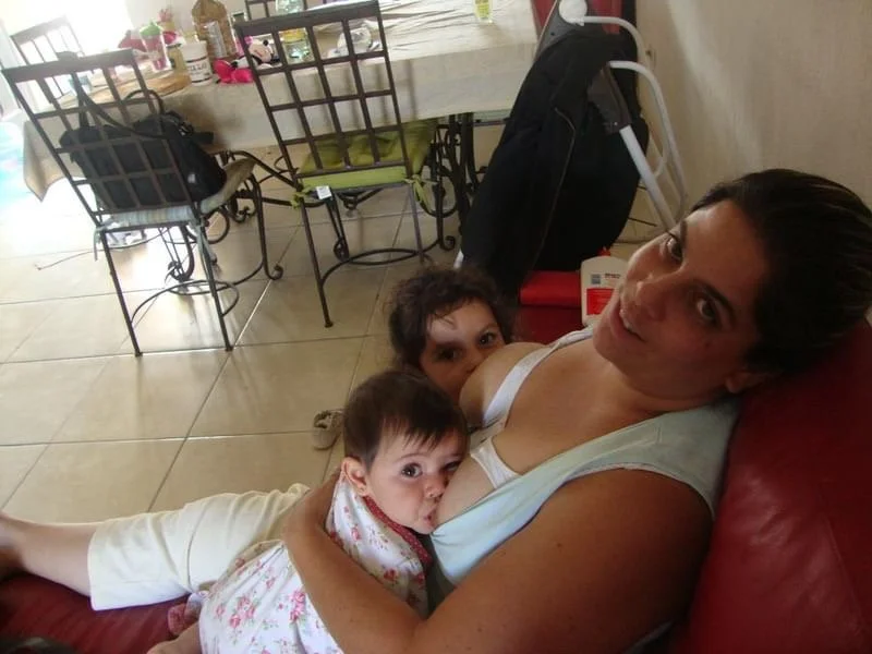 Mother breastfeeding two children, one toddler and one infant. They are sitting on a bench in the kitchen.