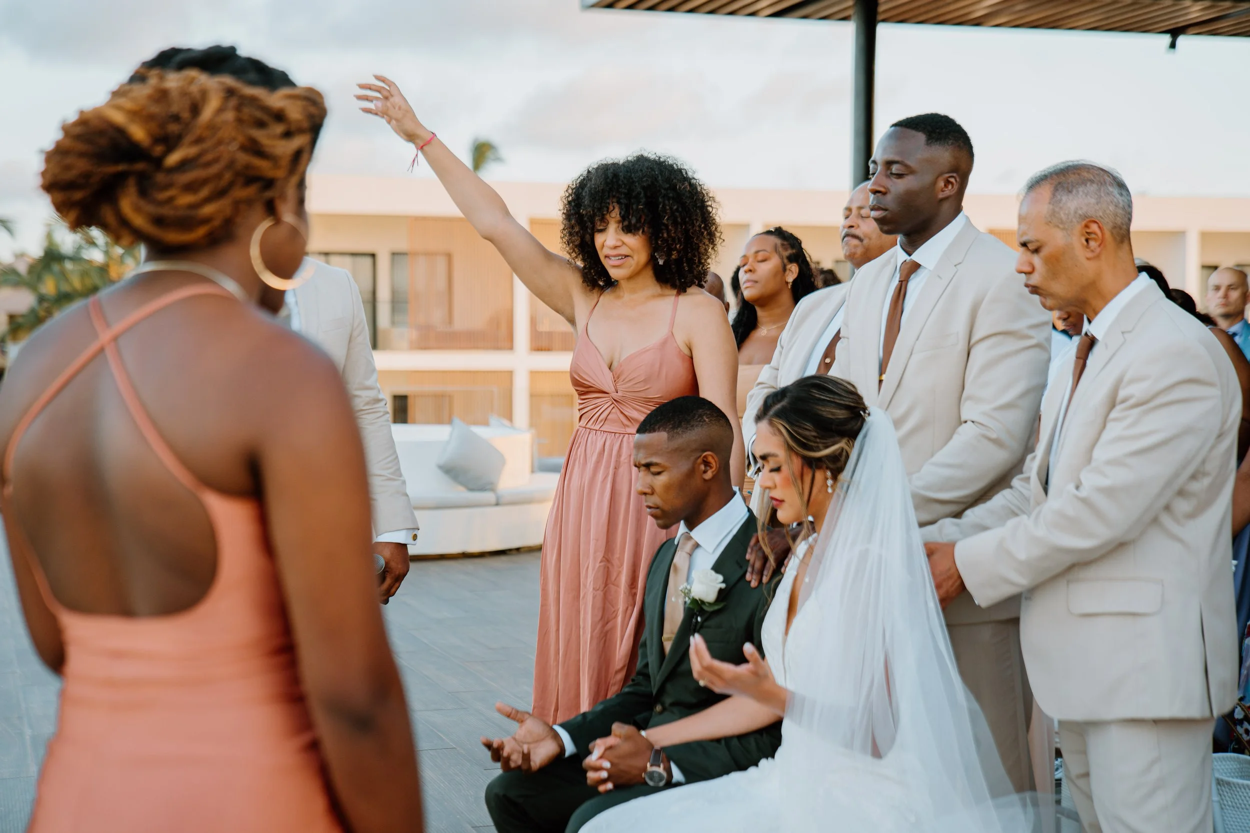 A wedding group participates in a prayer ceremony. The bride and groom kneel with eyes closed, hands open. Bridesmaids and groomsmen, mostly in beige attire, surround them with bowed heads and hands of blessing. The setting appears to be outdoors on 