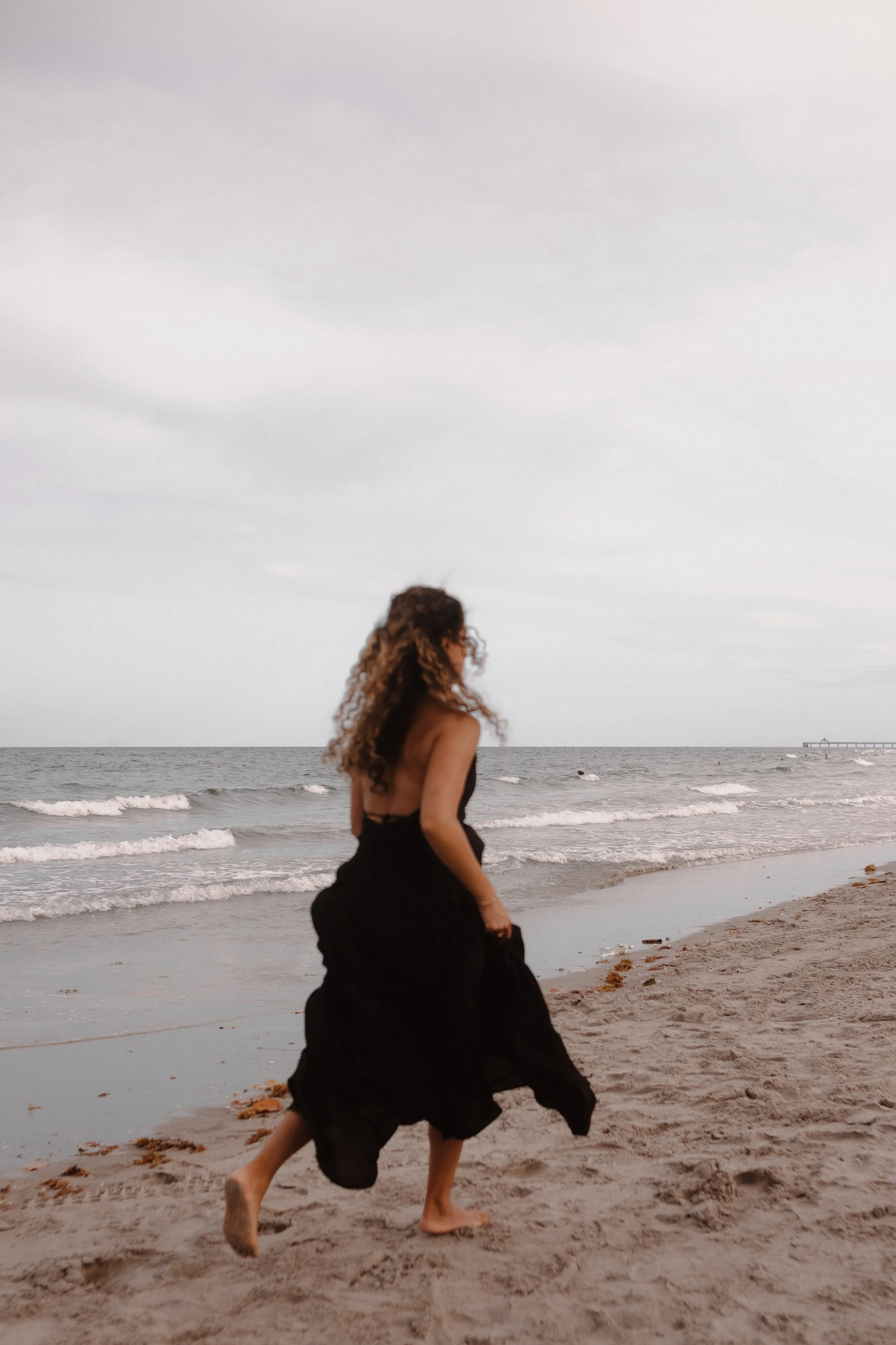Woman in black dress walking on a beach with waves in the background.