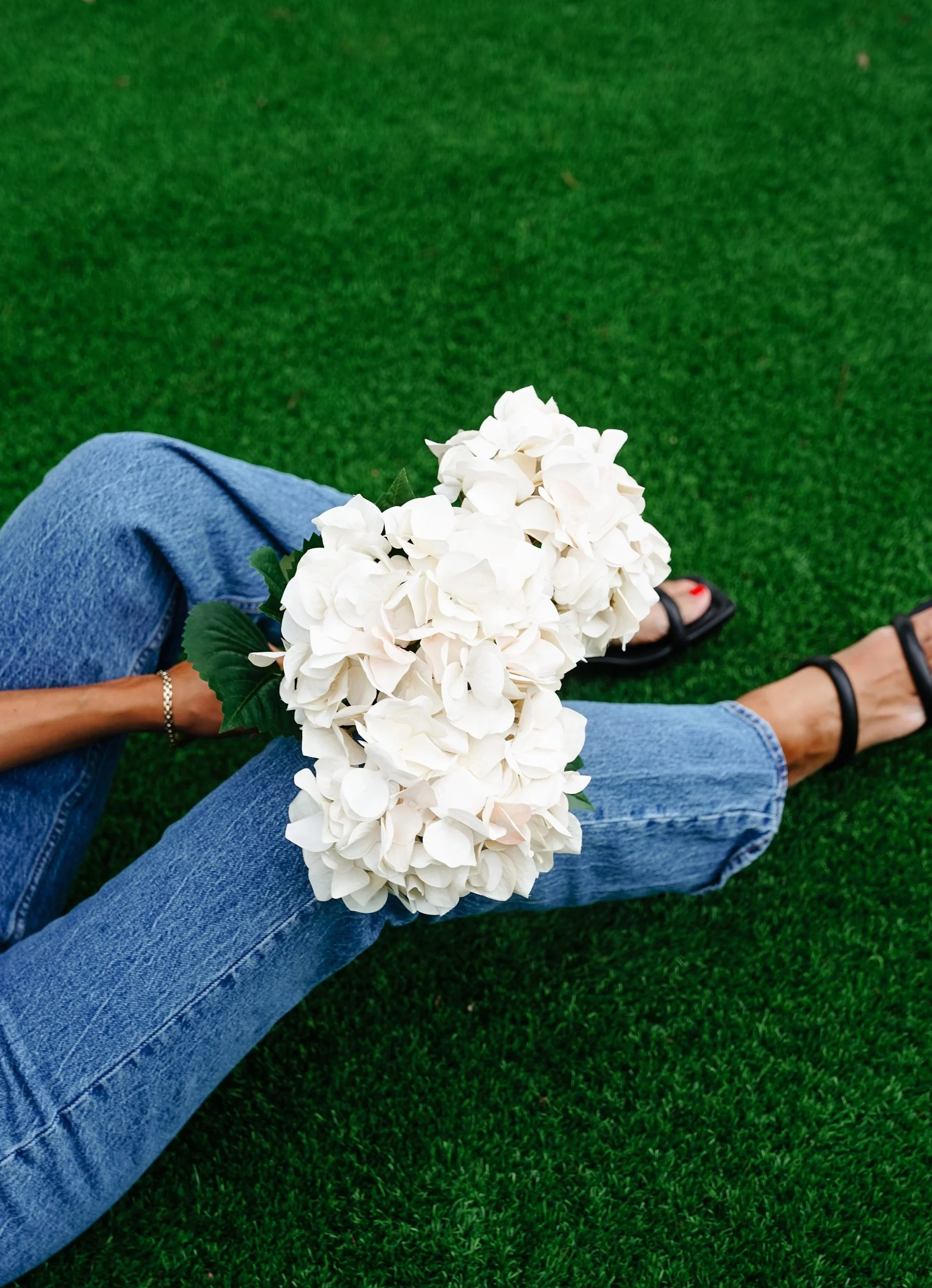 Person sitting on green grass, wearing blue jeans and black sandals, holding a bouquet of white hydrangea flowers.