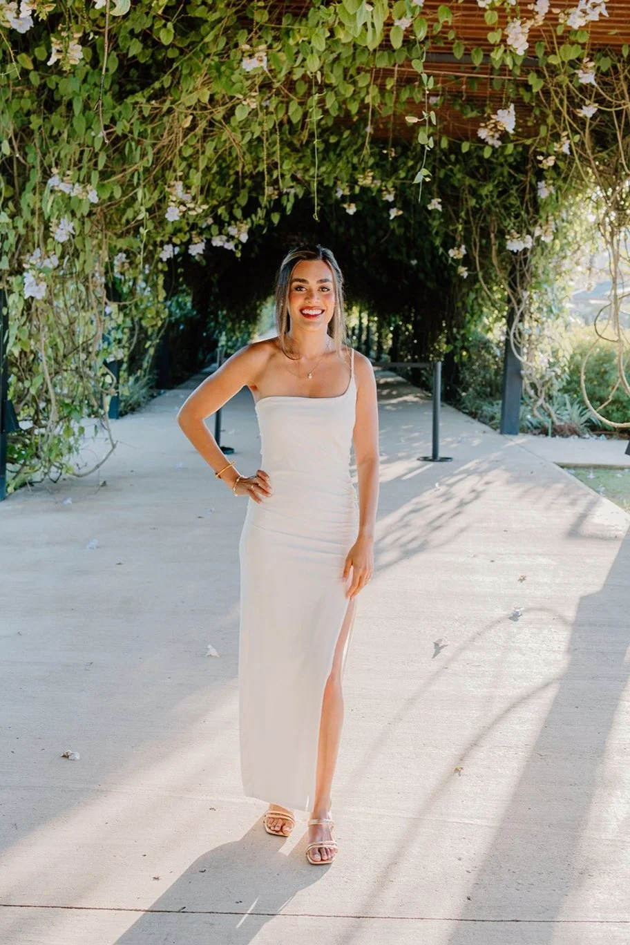 A woman in a white strapless dress with a slit walking outdoors on a sunny day, surrounded by greenery and flowering vines.