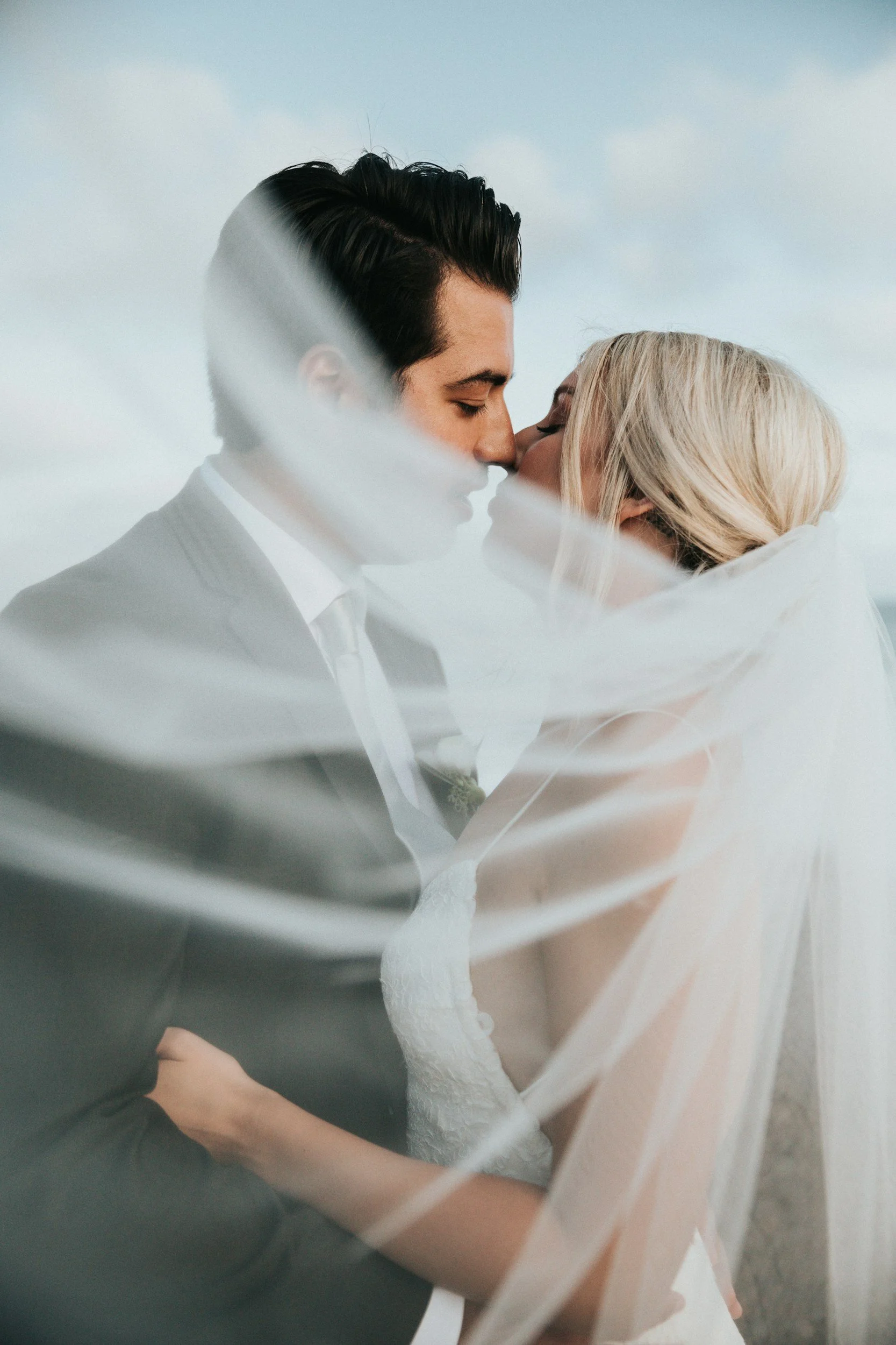 A man and a woman sharing a kiss, dressed in wedding attire, through a sheer fabric. The man has dark, styled hair and is wearing a suit, while the woman has long blonde hair and is wearing a wedding dress. The sky and clouds are visible in the background.