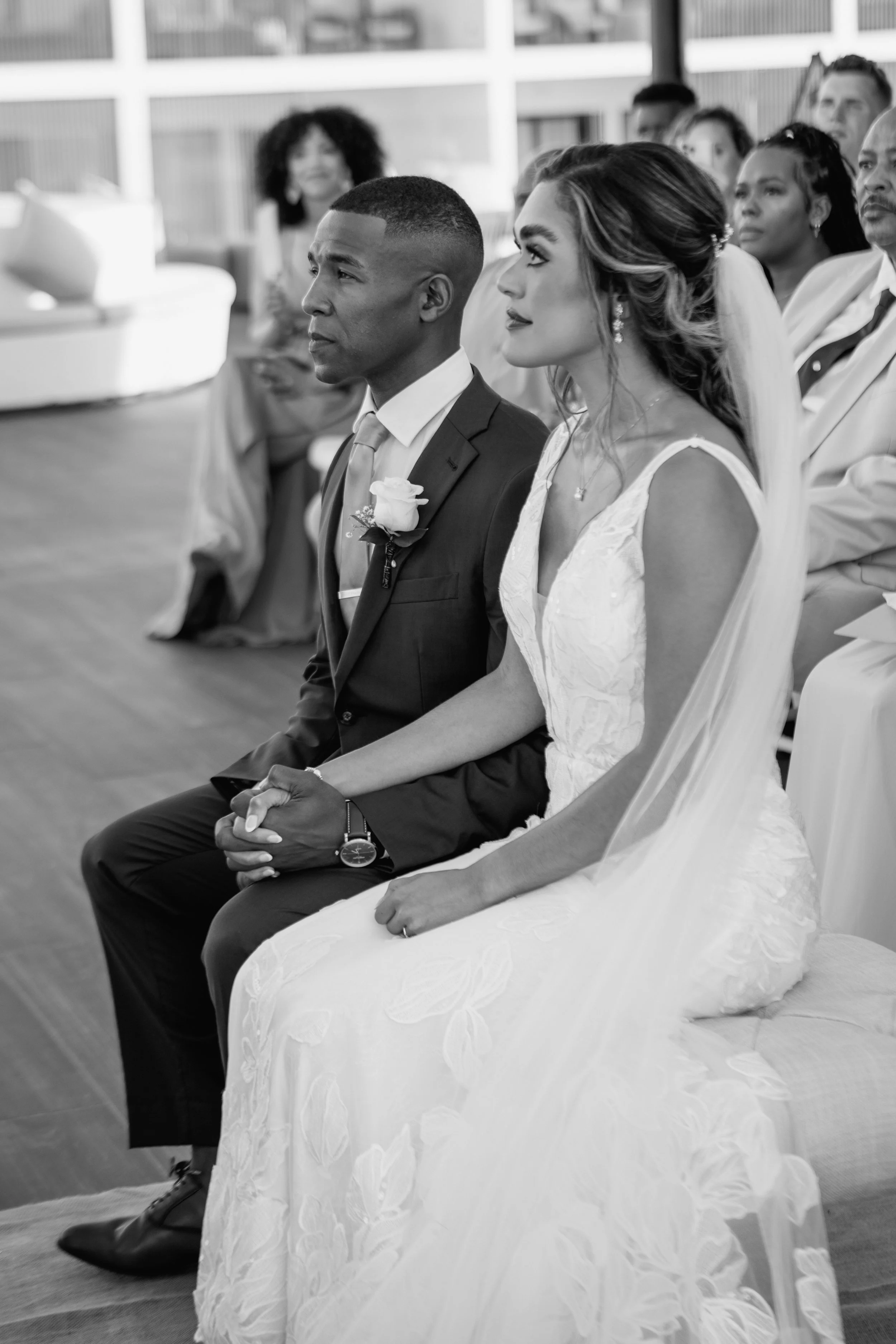Bride and groom sitting close, holding hands during wedding ceremony, surrounded by guests, black and white photograph.