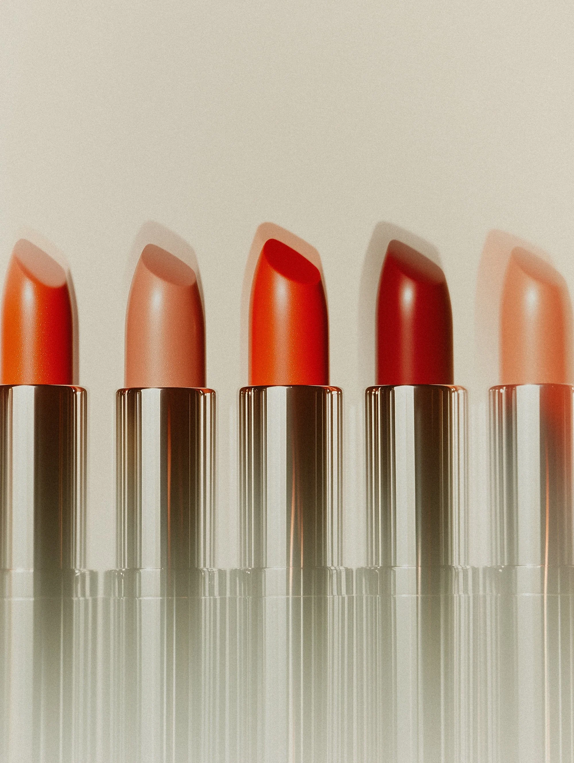 Close-up of six lipsticks in different shades of orange and red, arranged in a row against a plain neutral background.