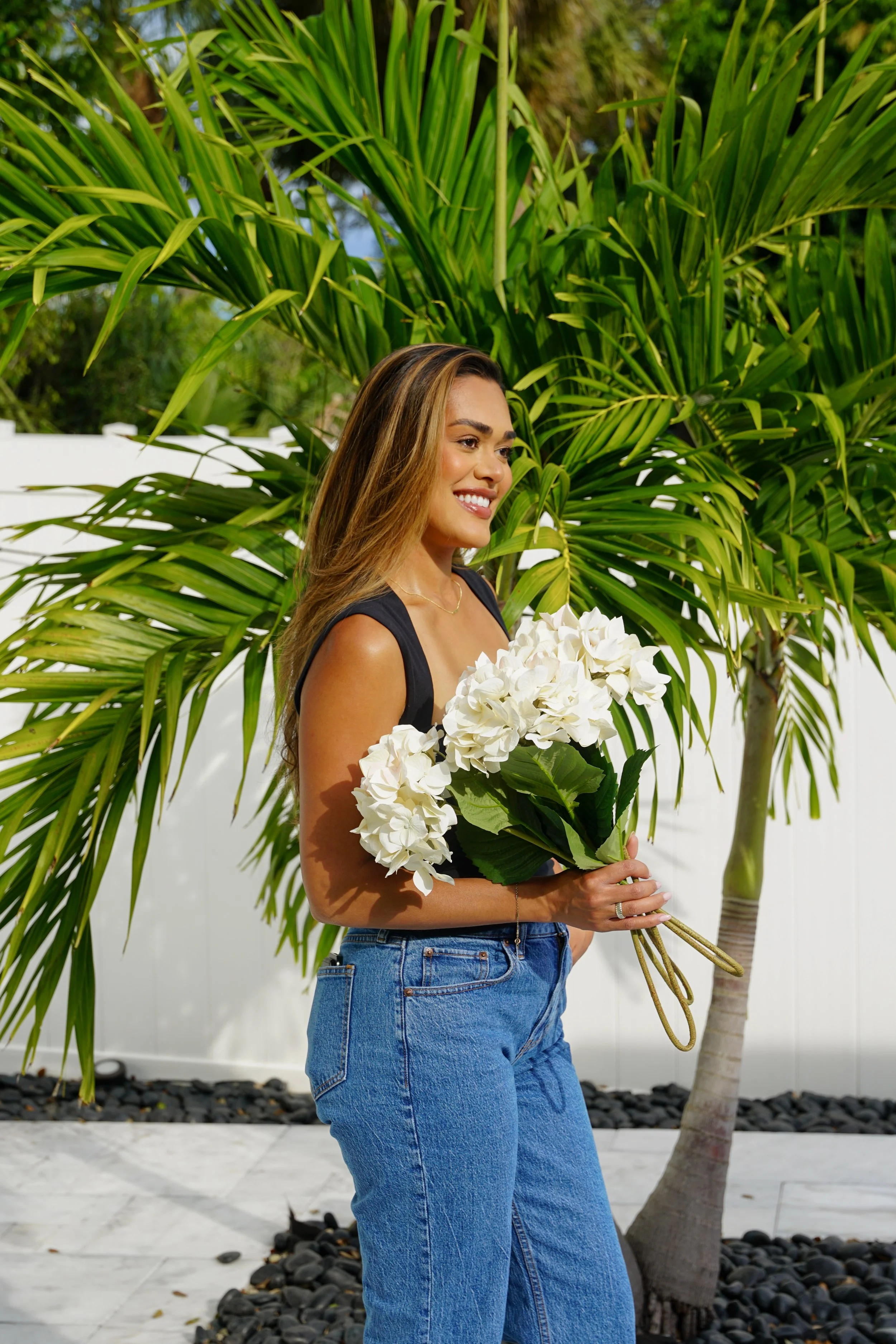 A woman in blue jeans and a sleeveless top holding a bouquet of white hydrangeas, standing outdoors near a green palm tree