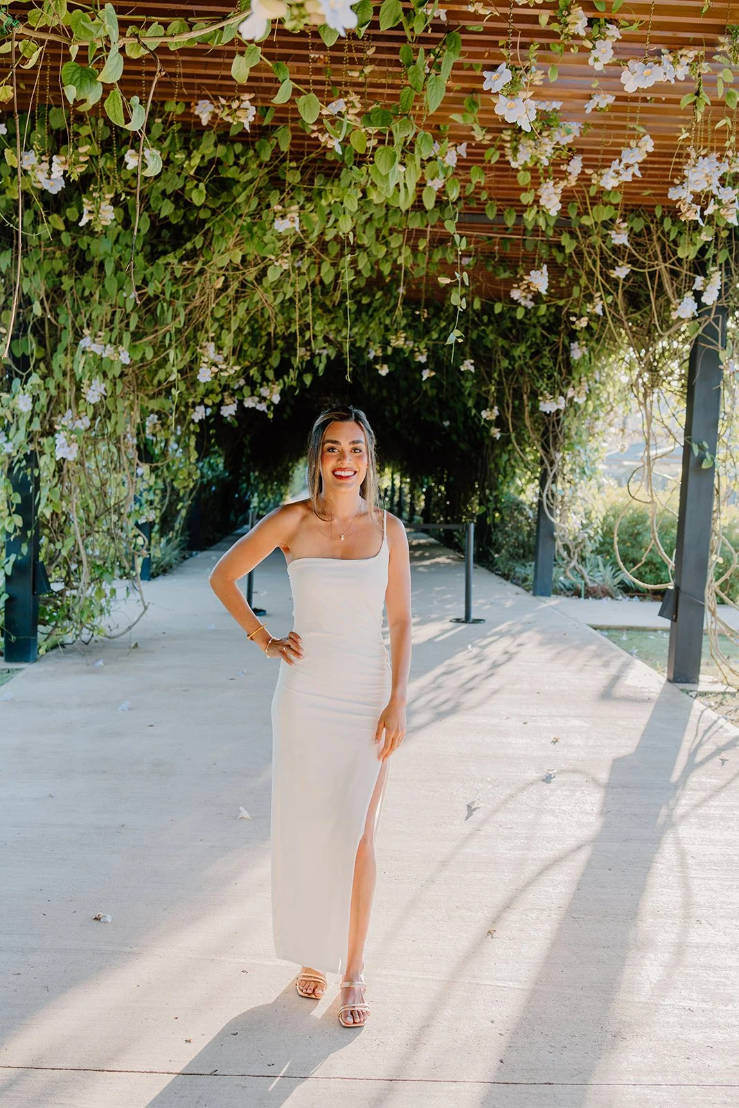 Woman in white dress standing under a floral vine-covered pergola.