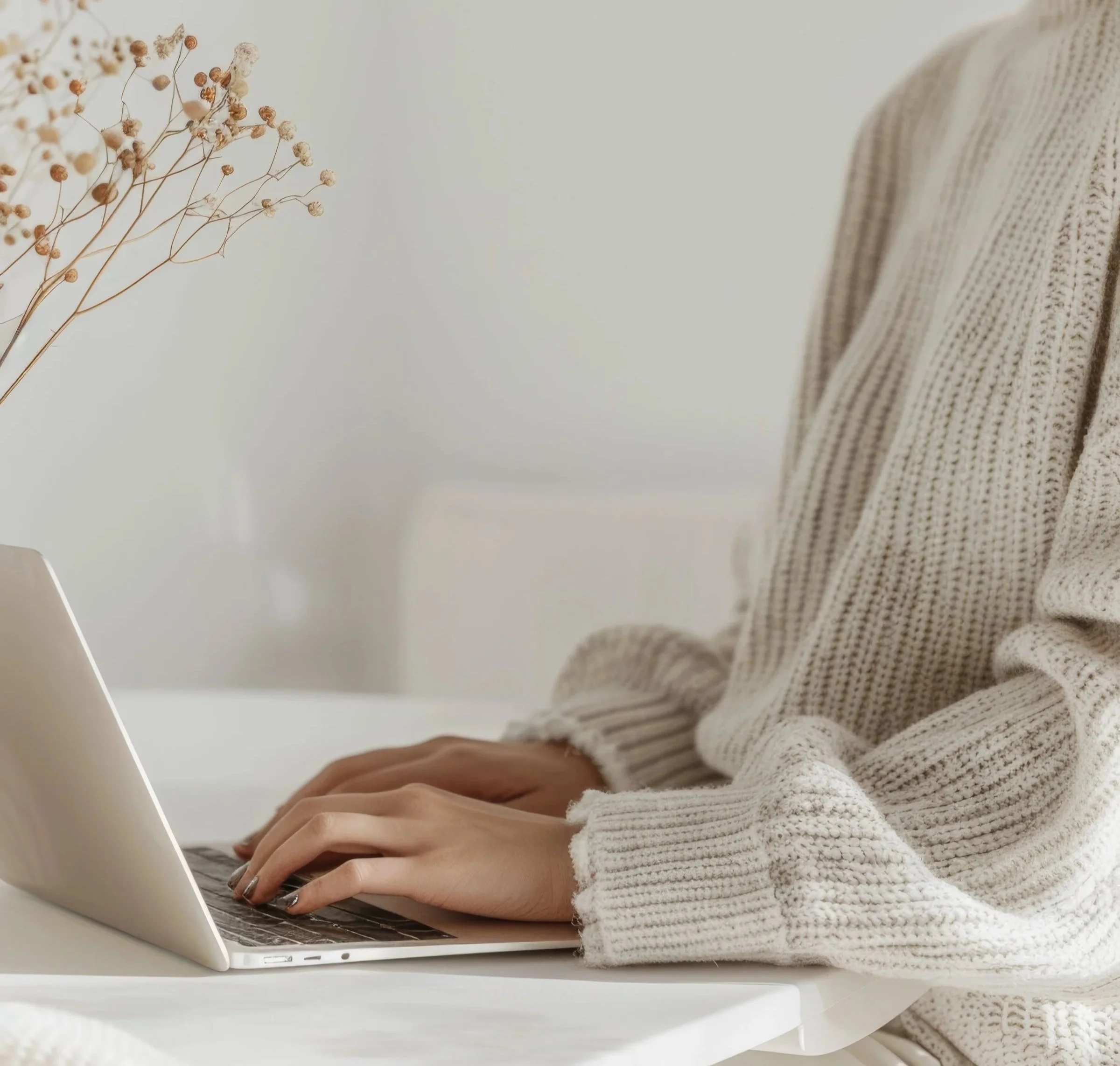 Person wearing a beige knitted sweater working on a laptop at a white desk, with a vase of dried flowers in the background.