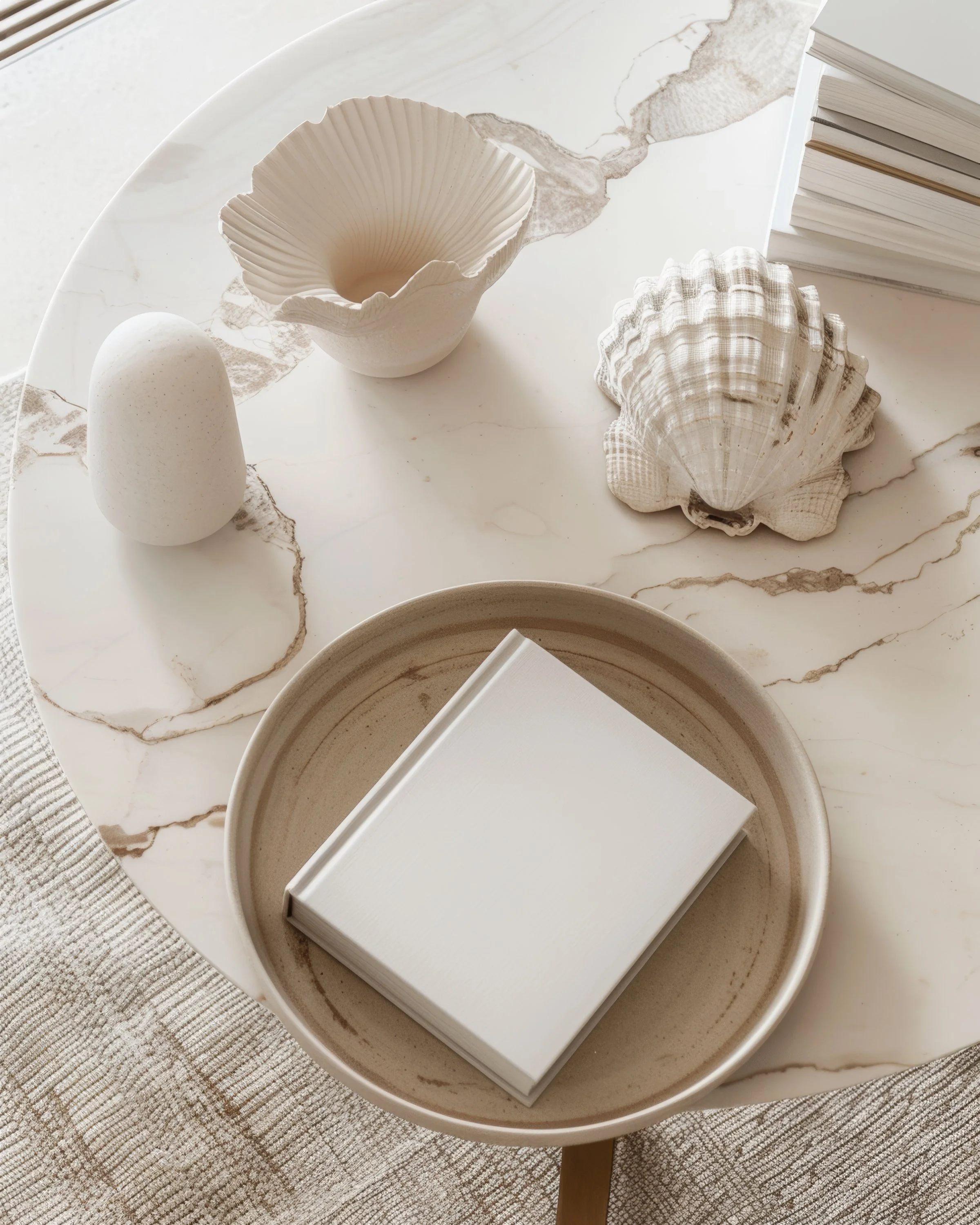 Decorative objects on a white marble table, including seashells, a white egg-shaped object, a small white bowl, a stack of white books, and a beige ceramic bowl with a closed white book inside.