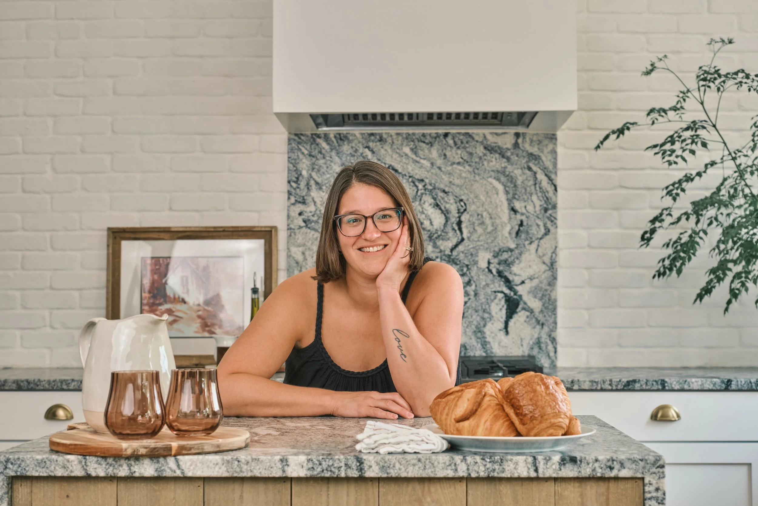 A smiling woman with glasses and shoulder-length brown hair sitting at a kitchen table. She is resting her head on her left hand, wearing a black sleeveless top. In front of her are three amber-colored drinking glasses, a white ceramic pitcher, and a plate of croissants. The background features a white brick wall with a framed picture, a granite backsplash, and a marble range hood above a stove. There is a large green plant on the right side.