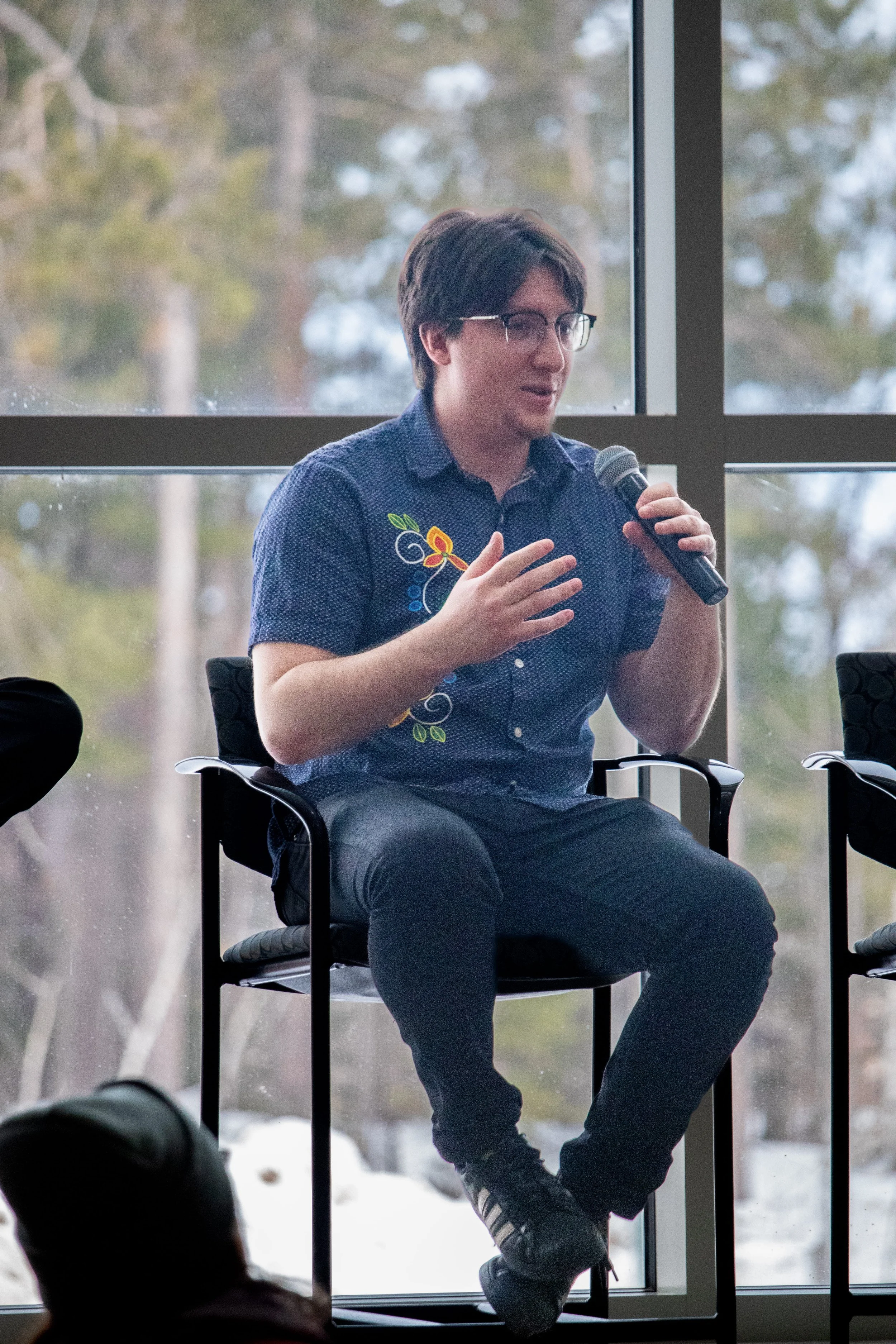 A person with short brown hair, glasses, wearing a dark blue shirt with colorful embroidery, is sitting in a chair and speaking into a microphone during a discussion or presentation. They are seated near large windows showing trees outside.