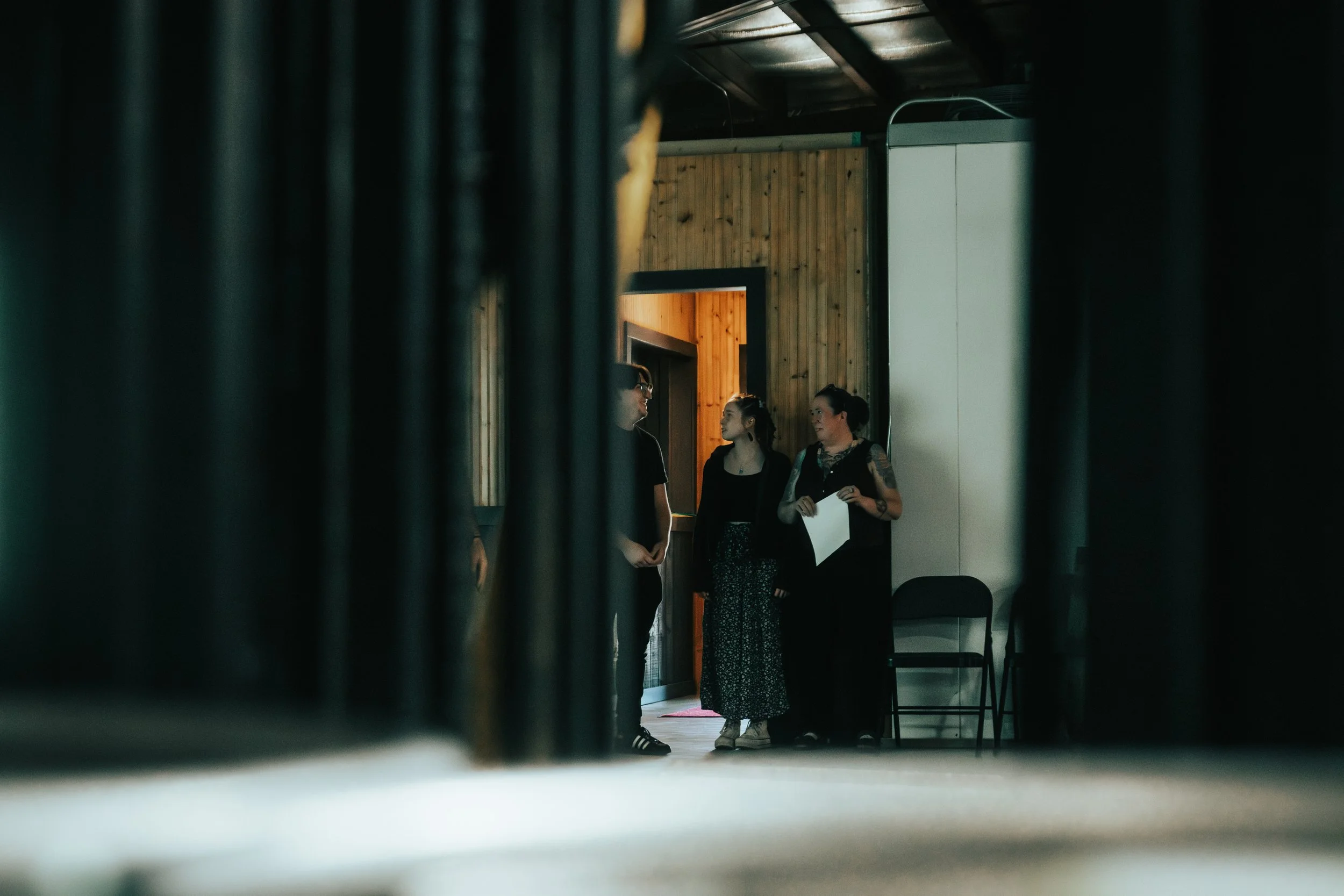 Three people standing in a room near a wooden wall, seen through a gap or narrow opening, with one person holding a piece of paper, having a conversation.